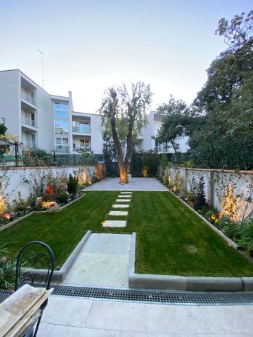 Cortile rettangolare ben illuminato con prato, sentiero in pietra e aiuole lungo un muro bianco, con condomini sullo sfondo.