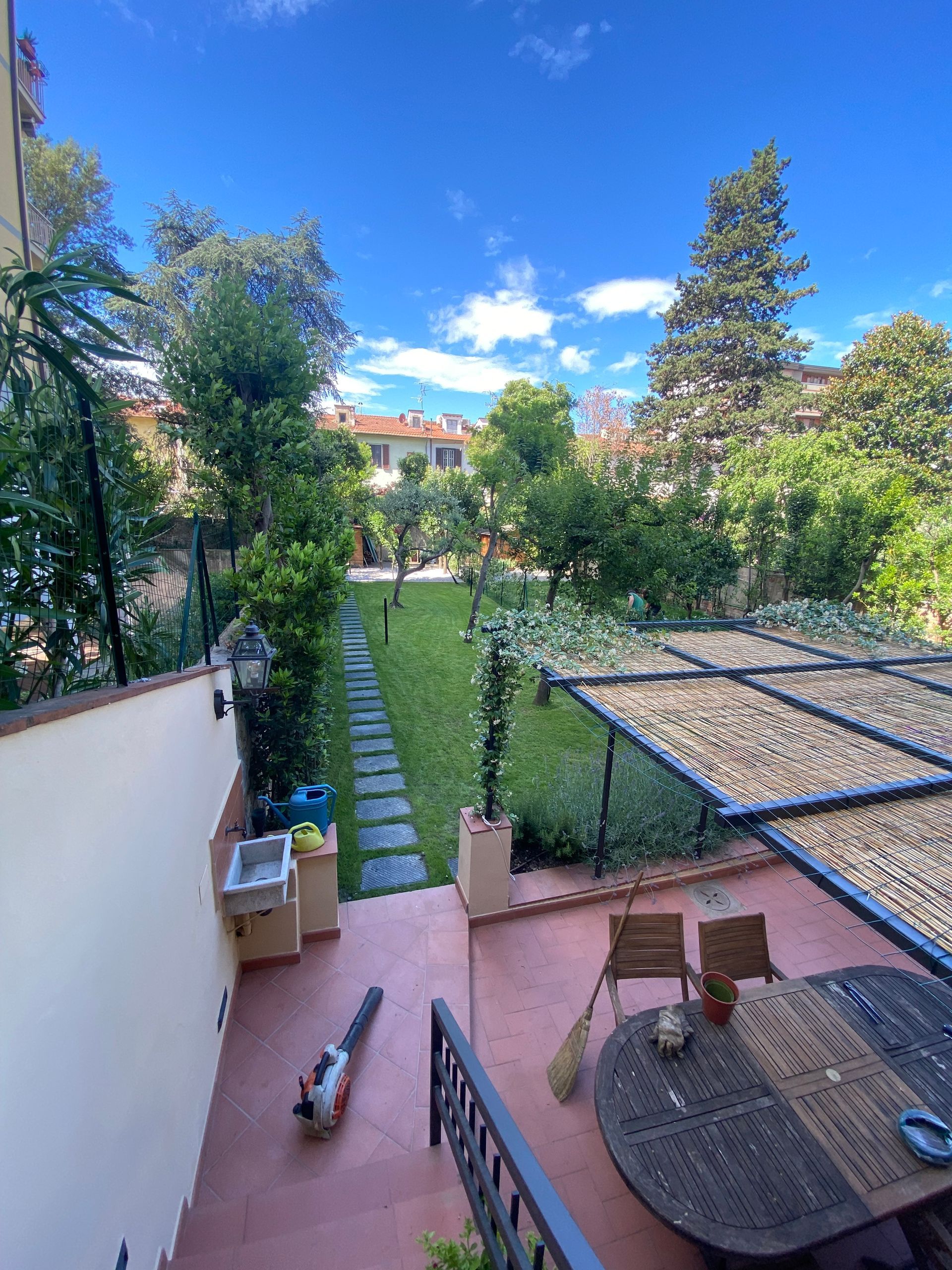 Giardino con patio, prato, alberi e cielo azzurro. Sono visibili un tavolo, delle sedie e un soffiatore per foglie.