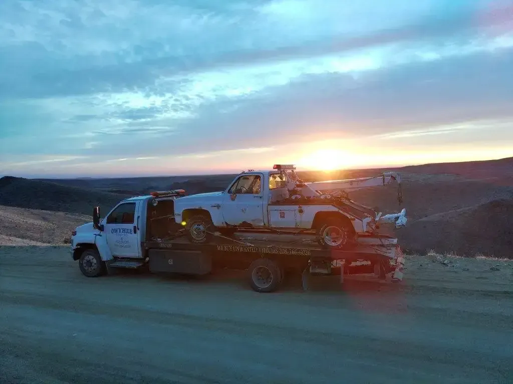 Tow truck hauling a white pickup truck on a dirt road at sunset.