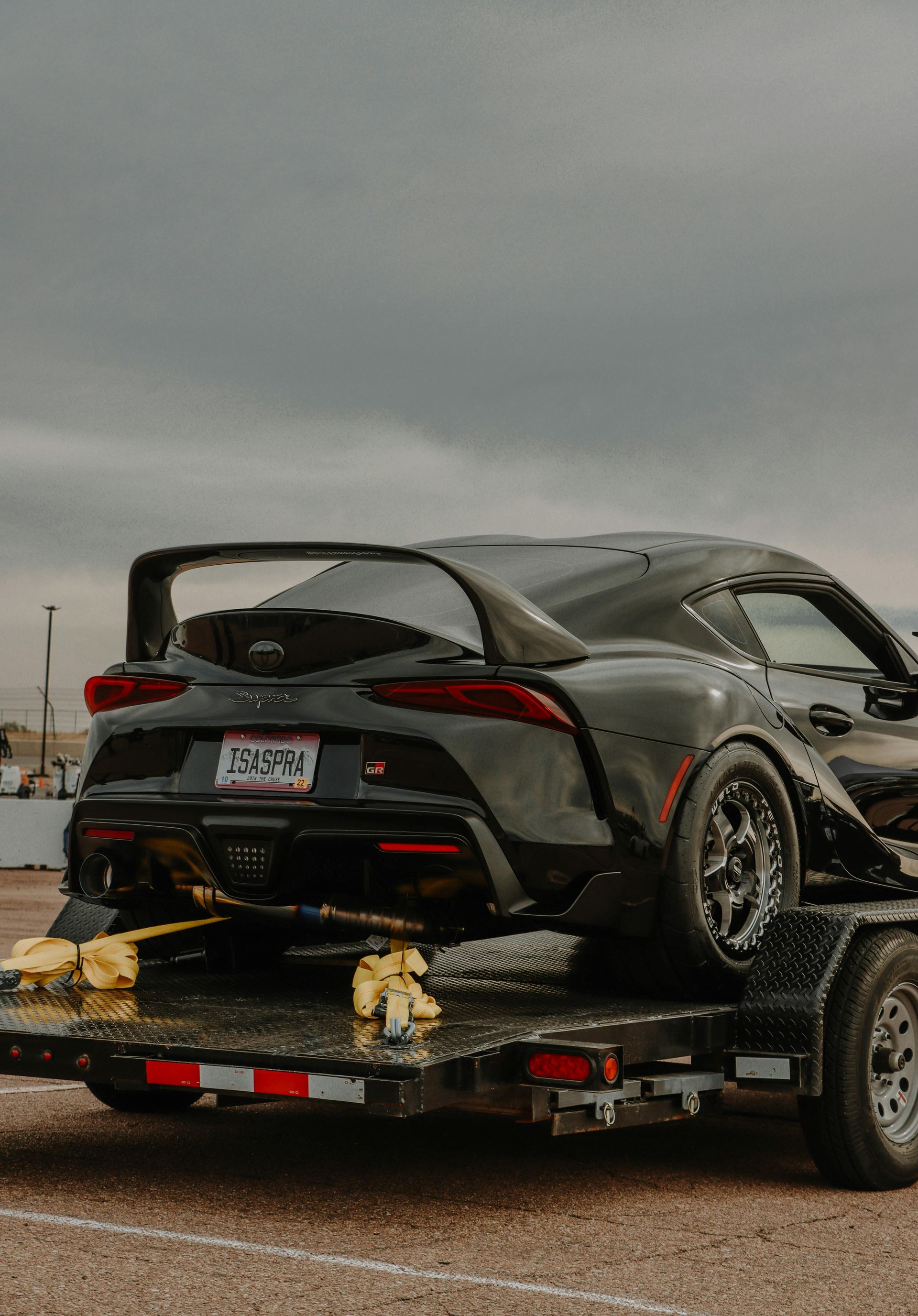 Black sports car on a trailer at a racetrack, under a cloudy sky.