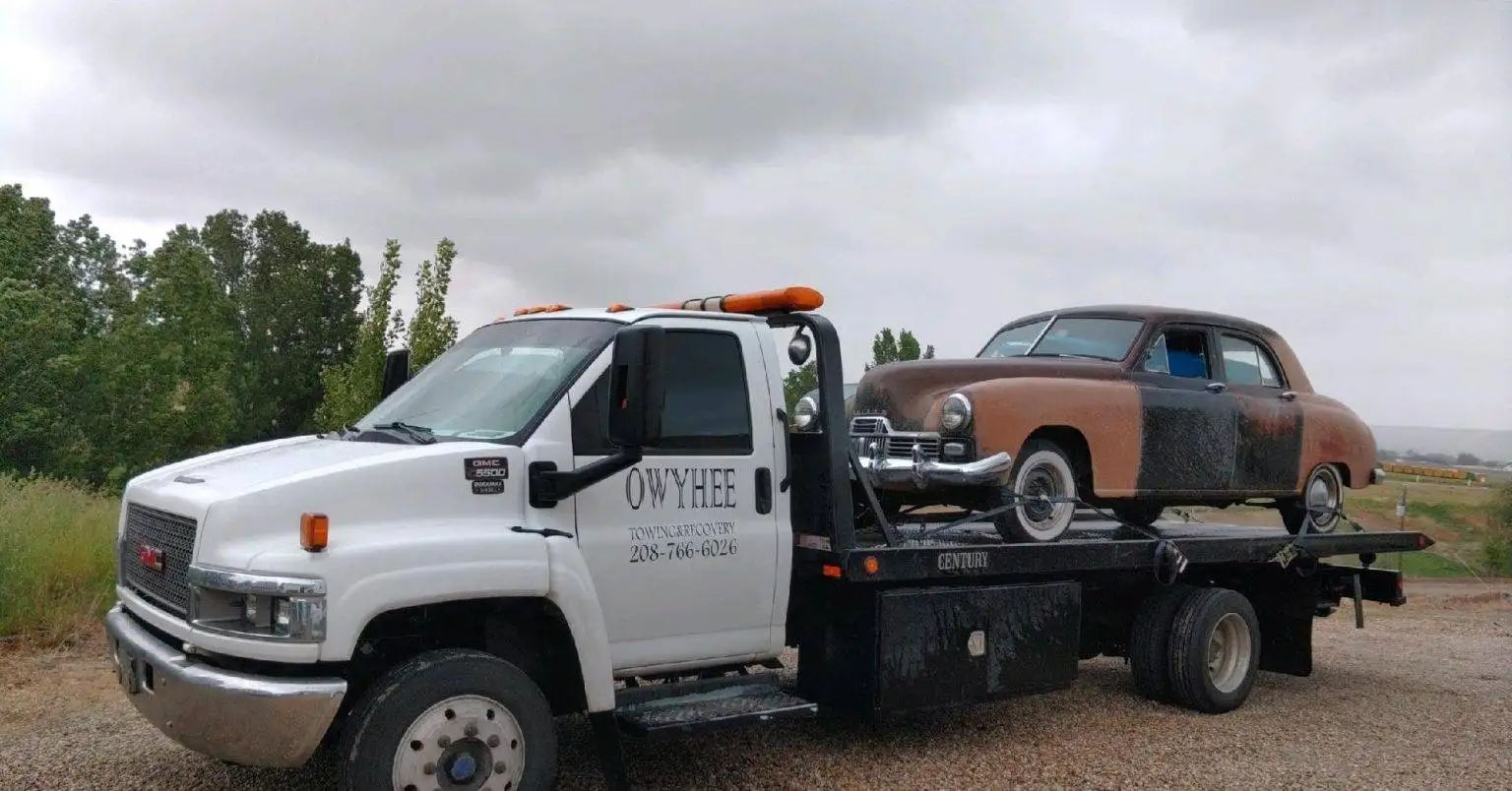 White tow truck hauling a vintage brown car on a cloudy day.