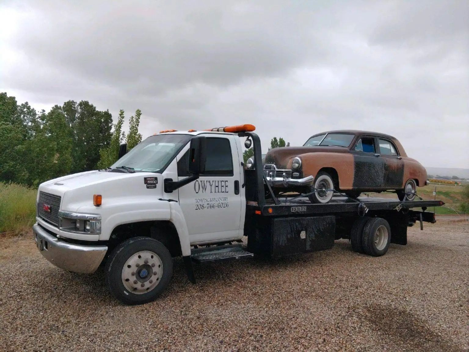 White tow truck with a vintage brown car on its flatbed, under a cloudy sky.