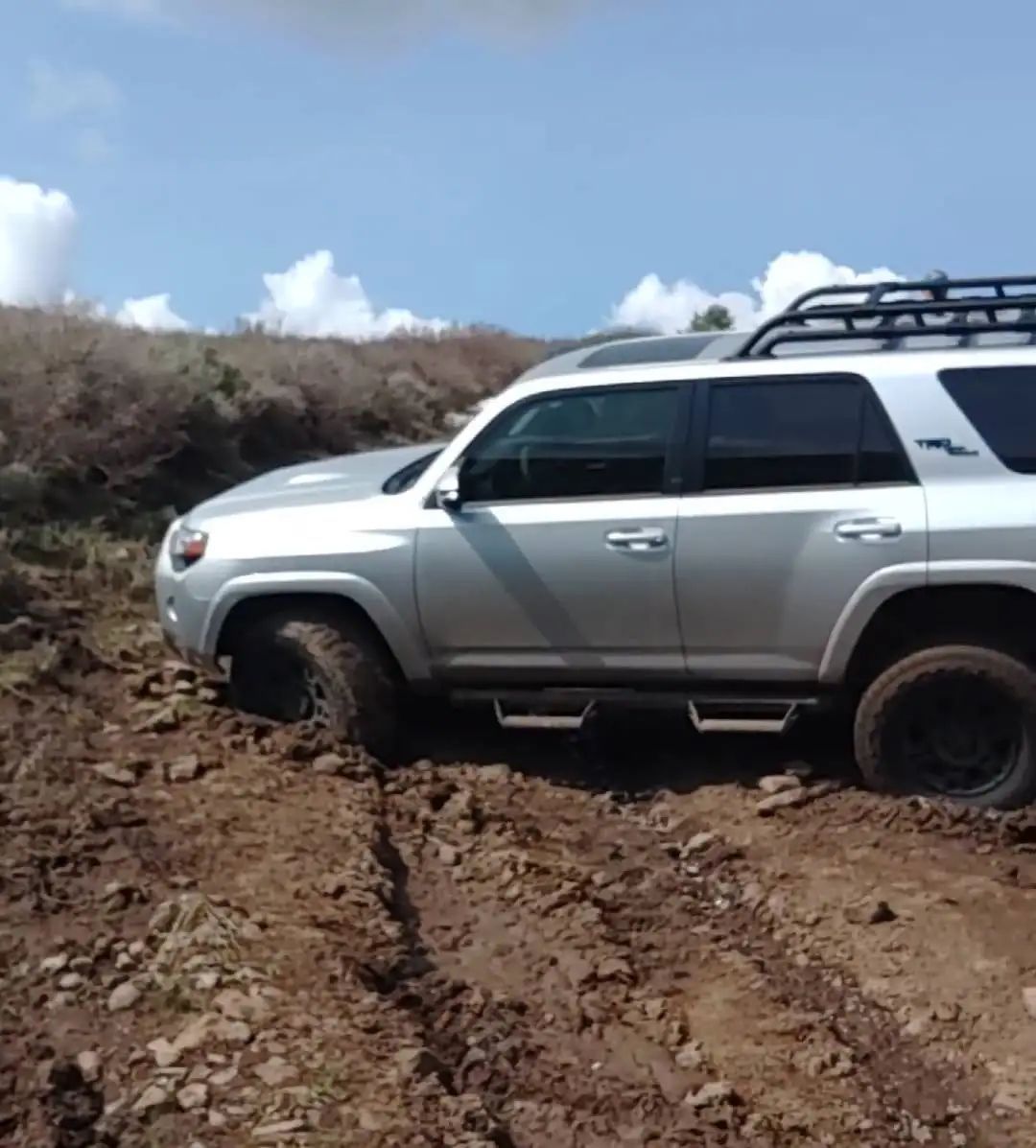 Silver SUV driving through muddy terrain on a sunny day.