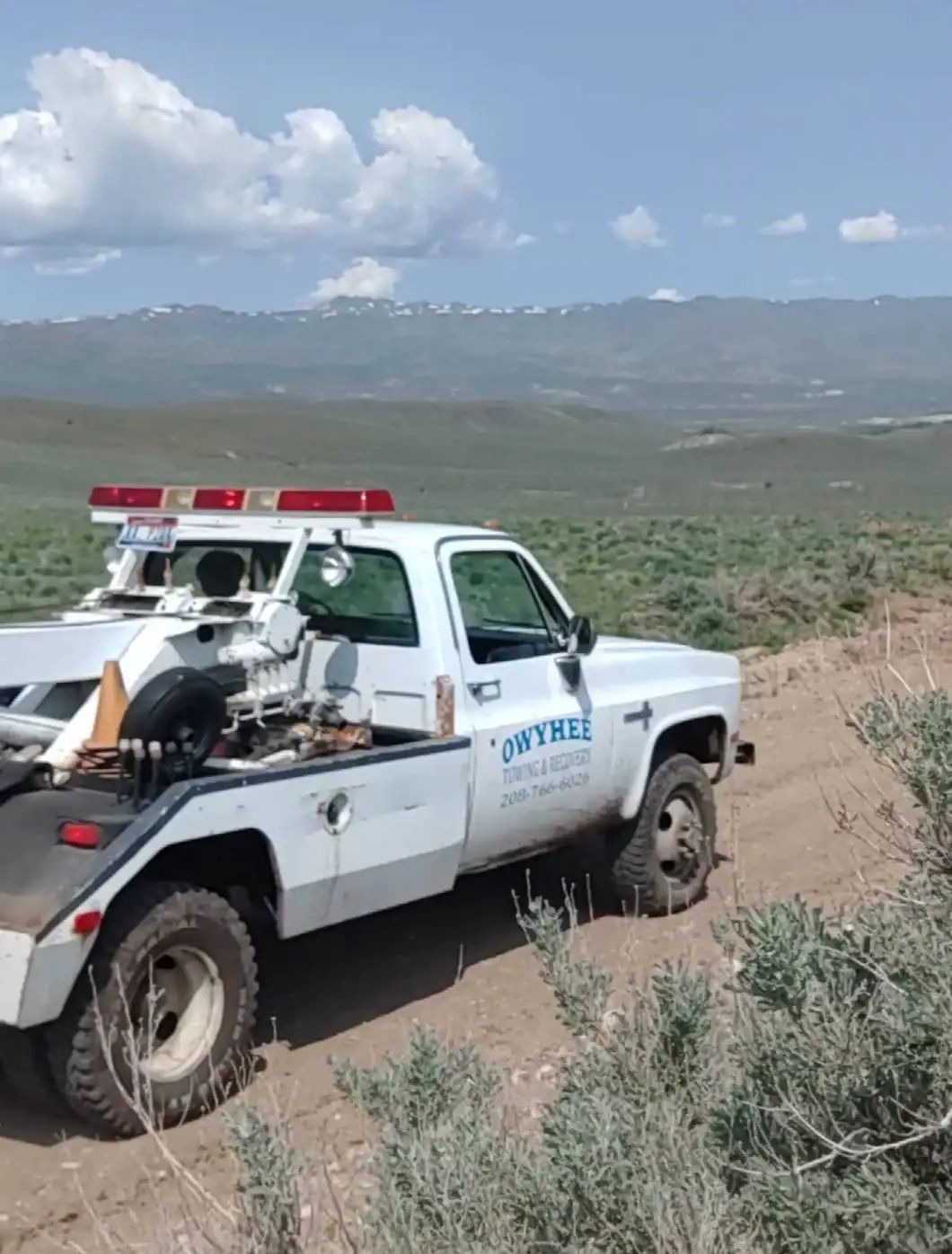 White tow truck on a dirt road, mountains in the background under a cloudy sky.
