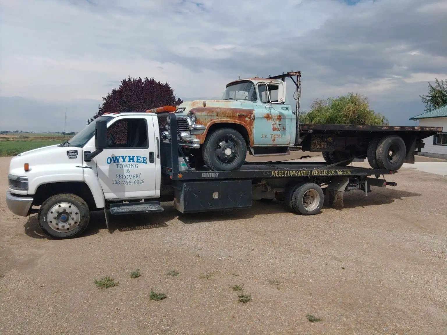 A rusty vintage pickup truck being transported on a tow truck in a rural setting.