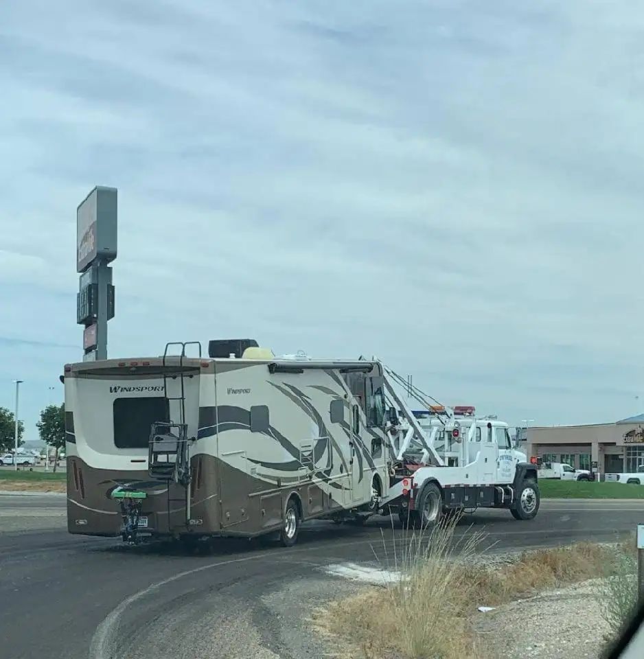 A white tow truck towing a large RV on a road, under a cloudy sky.