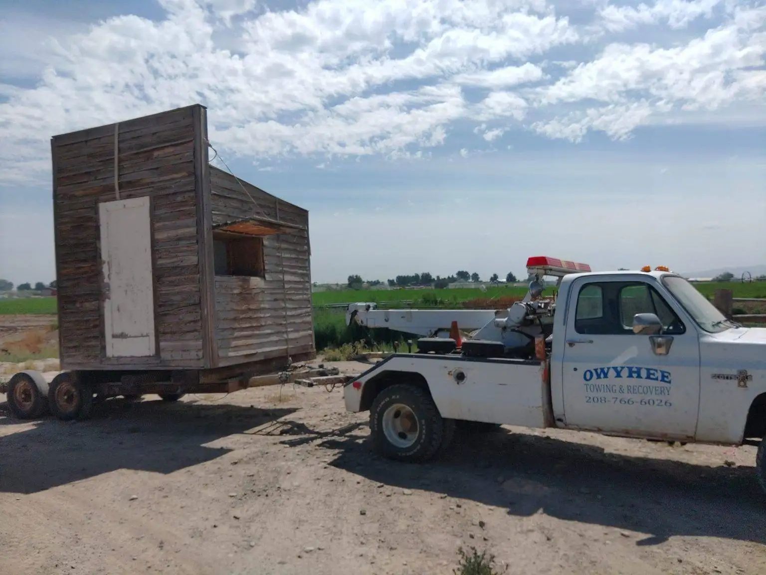 A tow truck pulling an old wooden structure on a trailer along a dirt road under a cloudy sky.