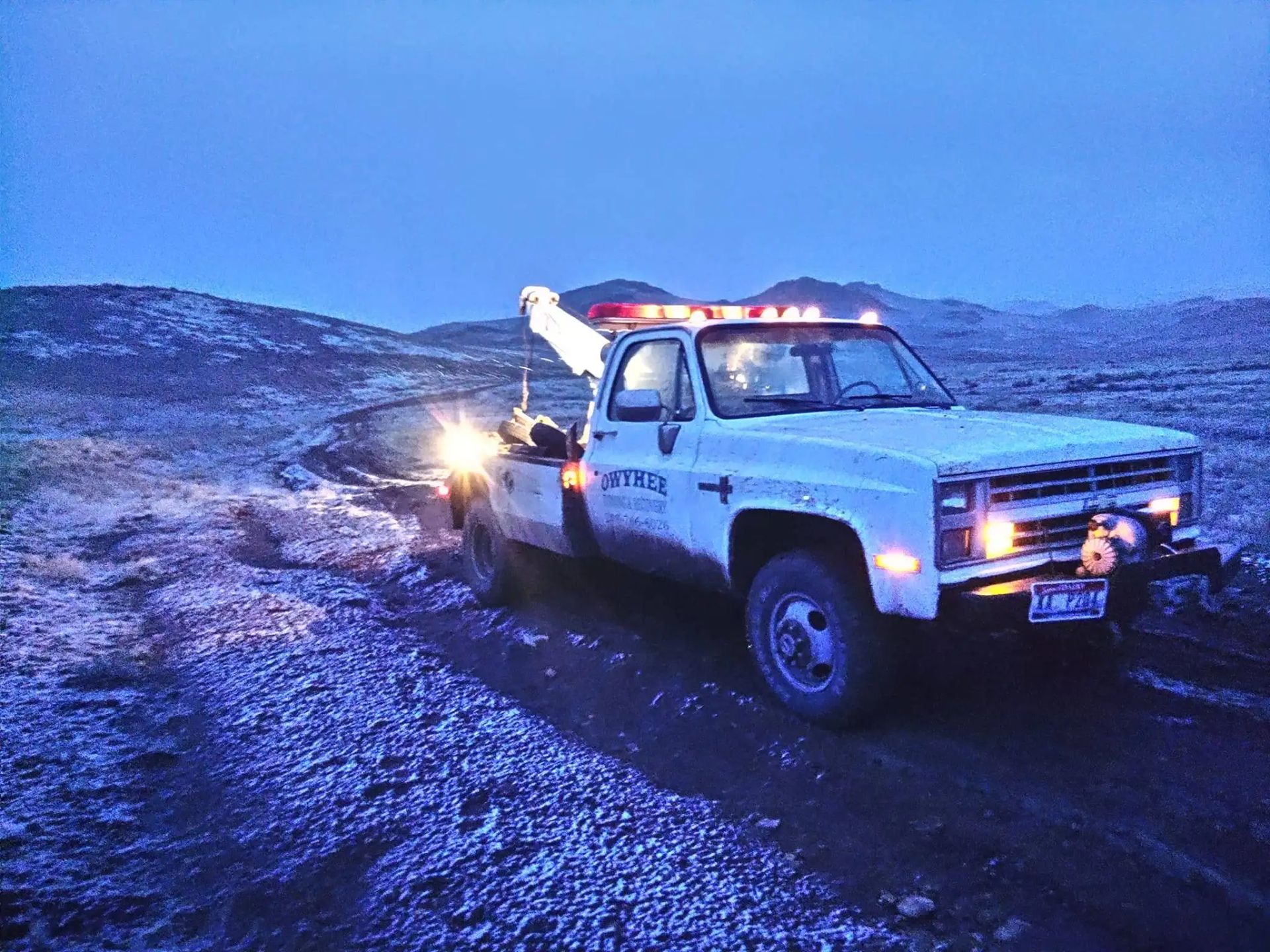Tow truck on a dirt road at dusk, lights illuminated, mountains in background.