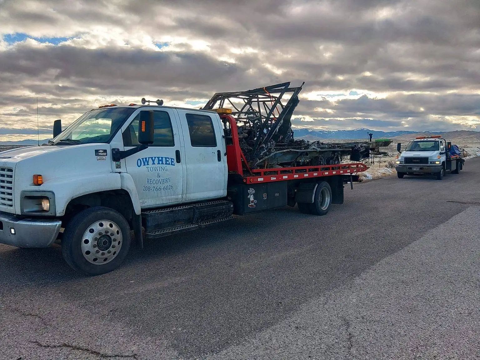 Two white tow trucks on a paved road under cloudy skies.