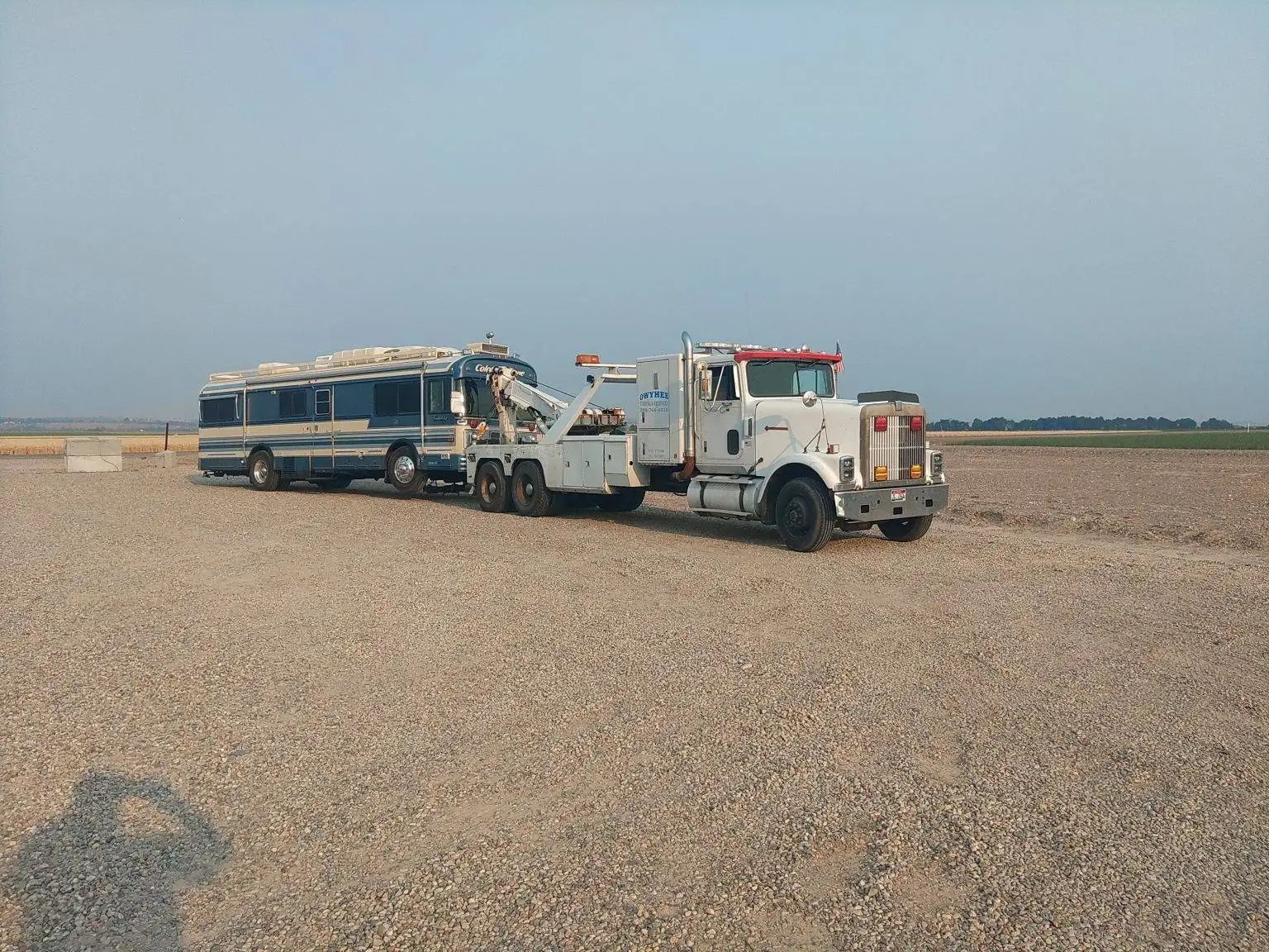 White tow truck towing a blue bus on a gravel surface under a blue sky.