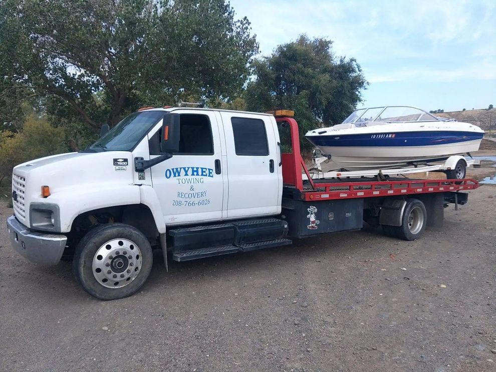 White tow truck transporting a boat on a trailer, outdoors.