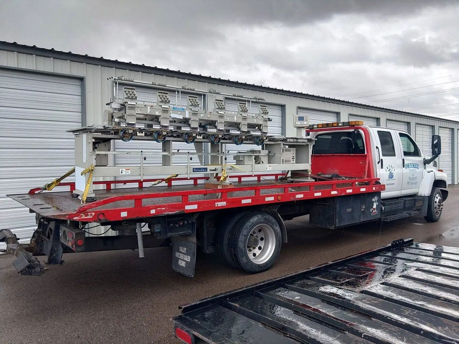 Tow truck hauling a large, silver industrial piece on a flatbed, parked near storage units.