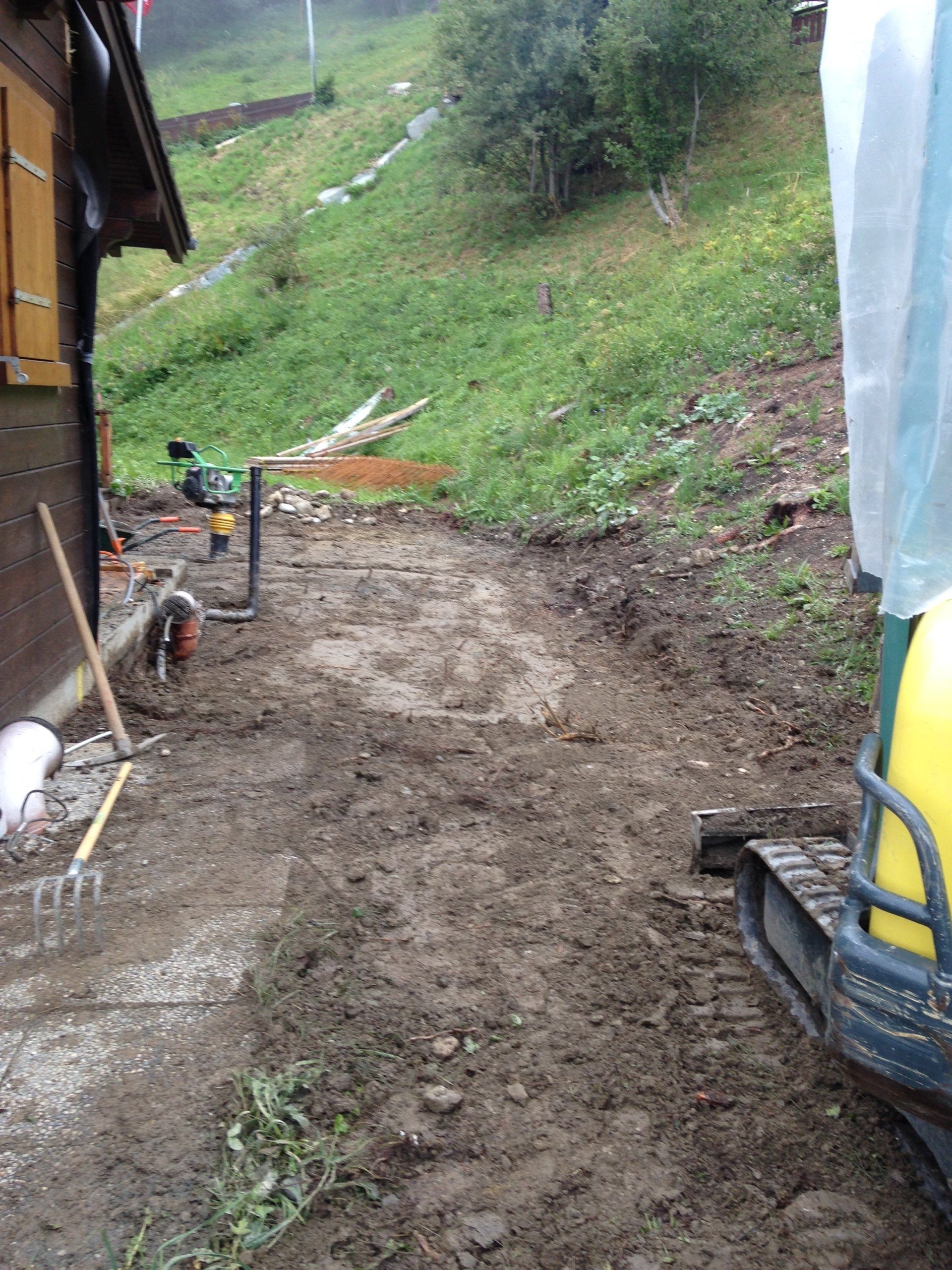 Une excavatrice jaune roule sur un chemin de terre à côté d'une maison.