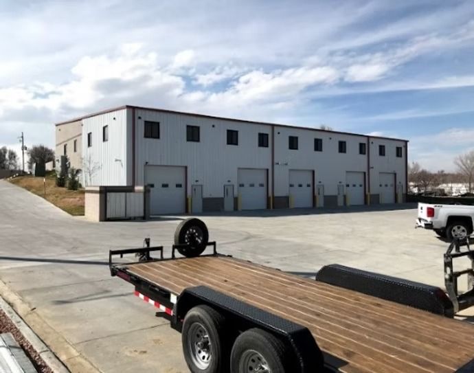A light-colored industrial building with multiple garage doors under a cloudy sky, a trailer in the foreground.