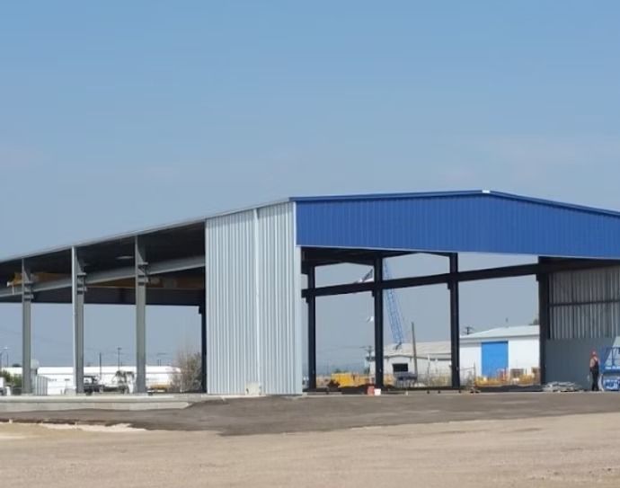 Blue-roofed metal building with open sides, likely a warehouse or storage structure, set on a gravel surface.
