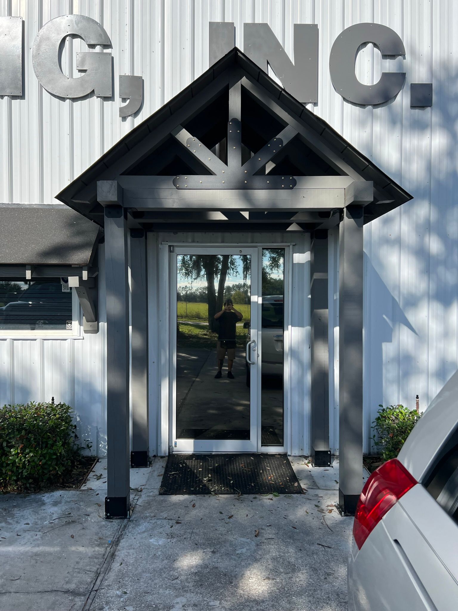 Gray-framed glass door with a small porch; building exterior; partially obscured person reflected in door.