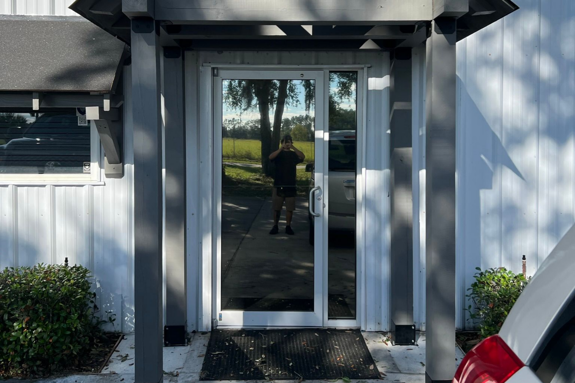 Person reflected in glass door, standing in front of building entrance, with a tree visible in the background.