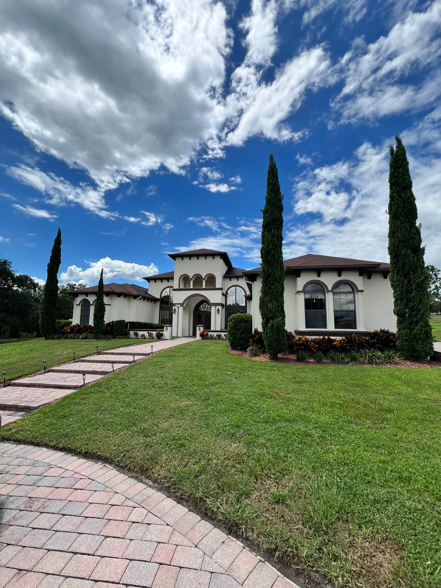 White stucco house with red tile roof, tall cypress trees, and brick walkway on a sunny day.