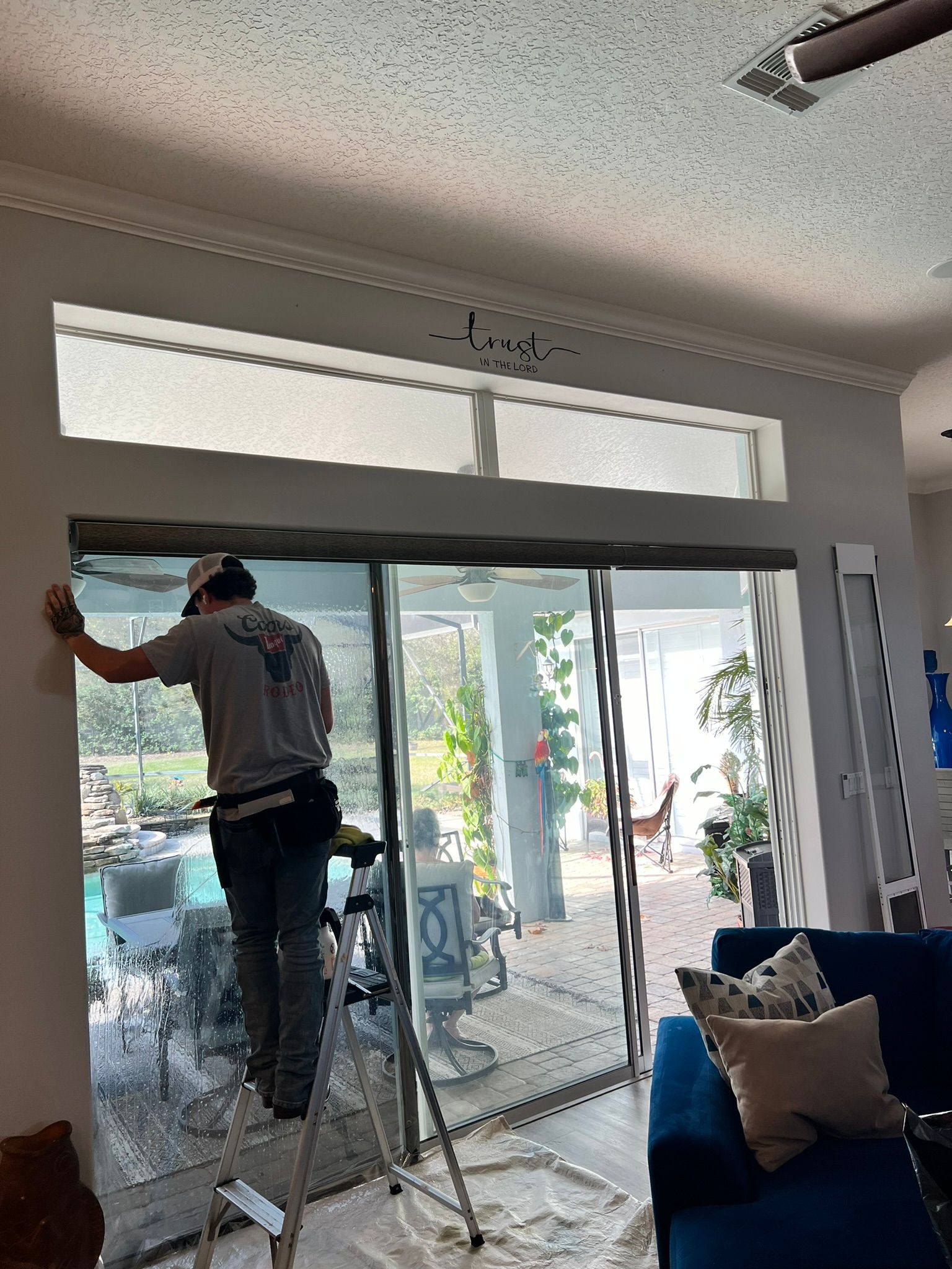 Man on ladder installing window tint on sliding glass door, overlooking a pool and backyard.