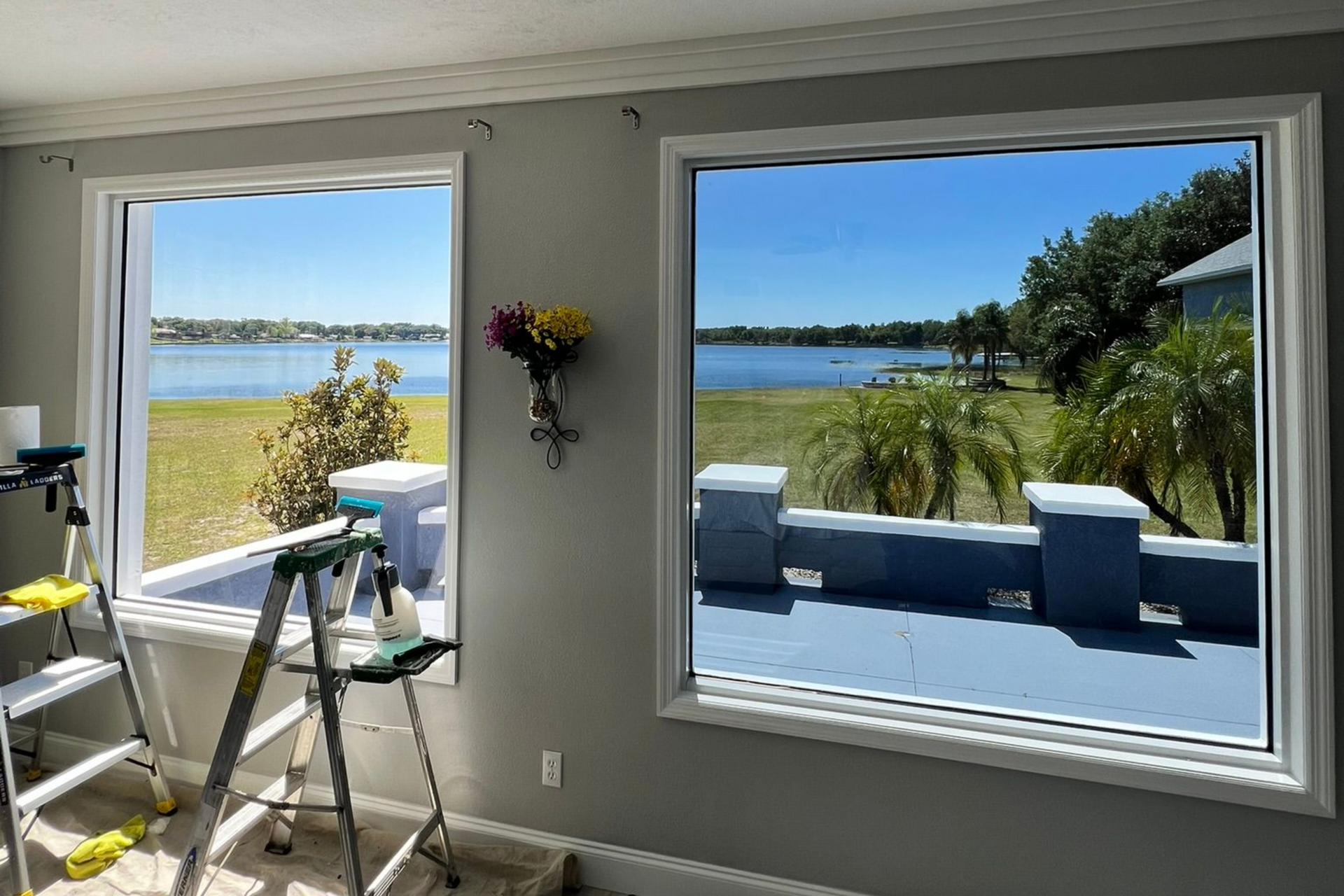 Two open windows with views of a lake and lawn, a ladder and cleaning supplies inside.