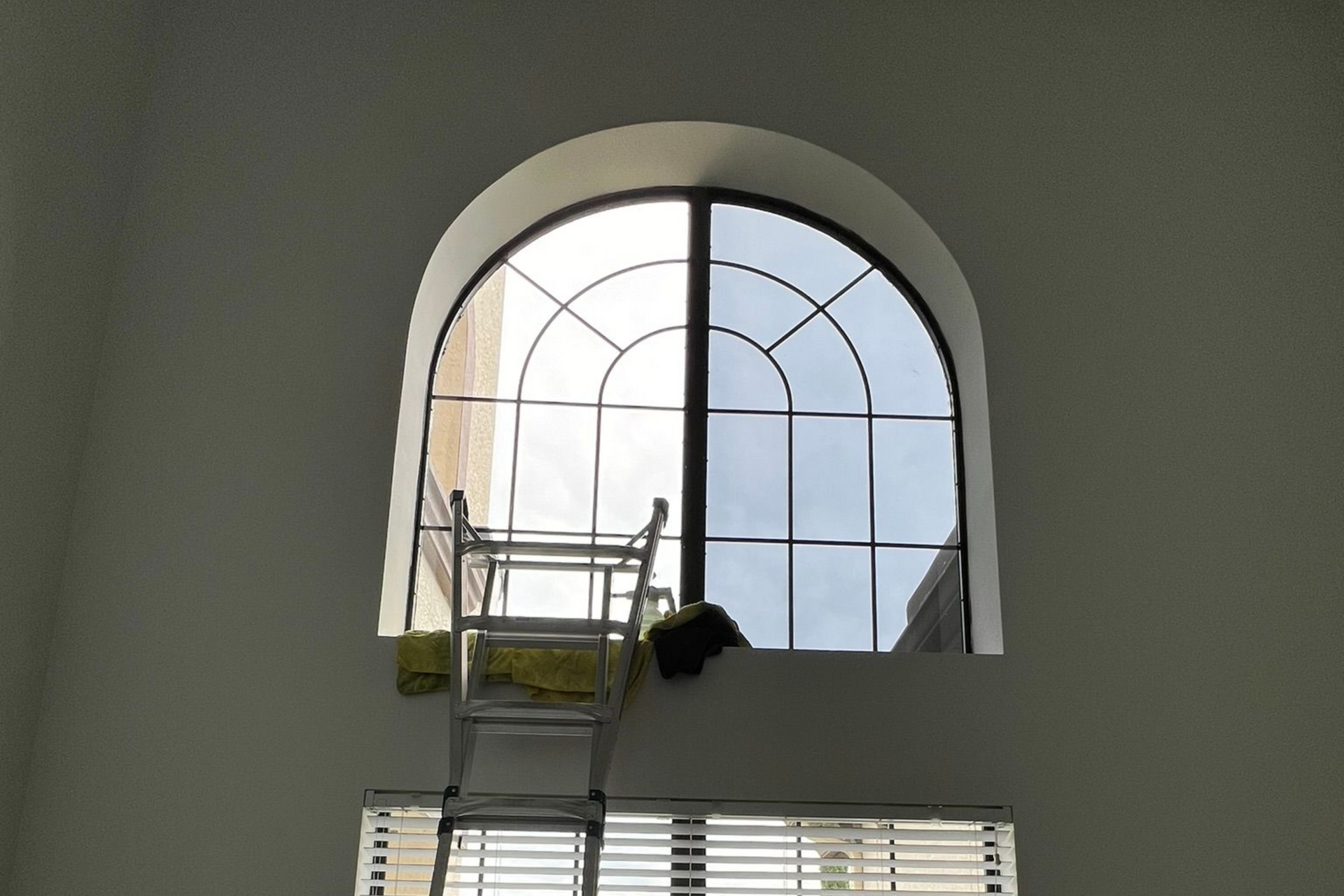 Arched window with black grid. Ladder inside, above blinds. Bright daylight shines through.