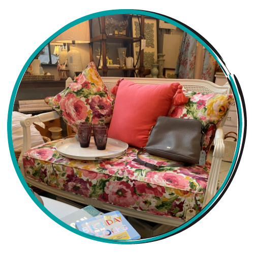 Floral-patterned wicker sofa with pink cushions, flowers, tray, and handbag in a cozy indoor setting