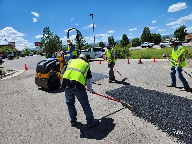 Workers paving a road with asphalt, using a roller and rakes, under a blue sky.
