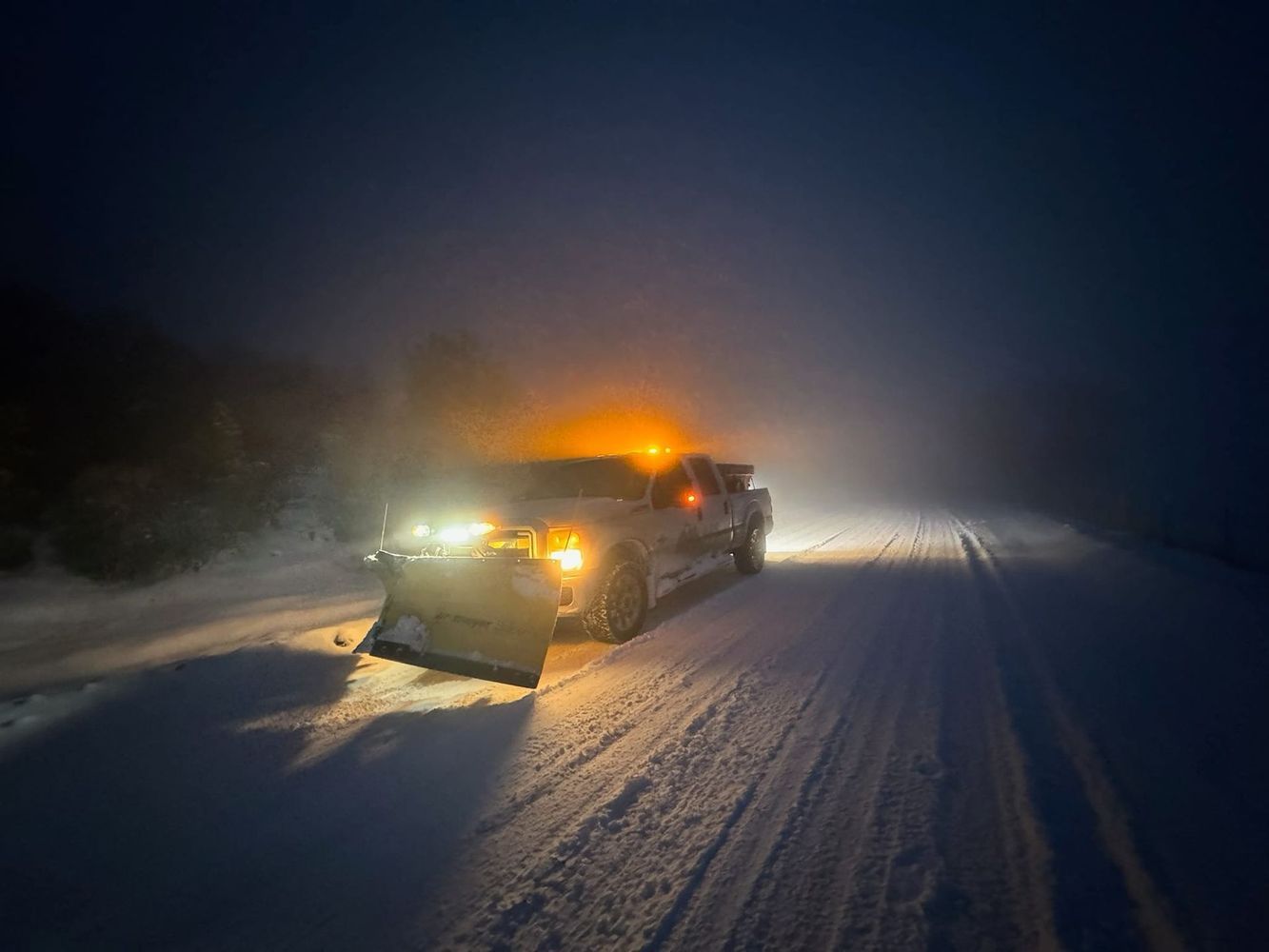 Snowplow clearing a snow-covered road at night, illuminated by headlights and orange hazard lights, with foggy conditions.