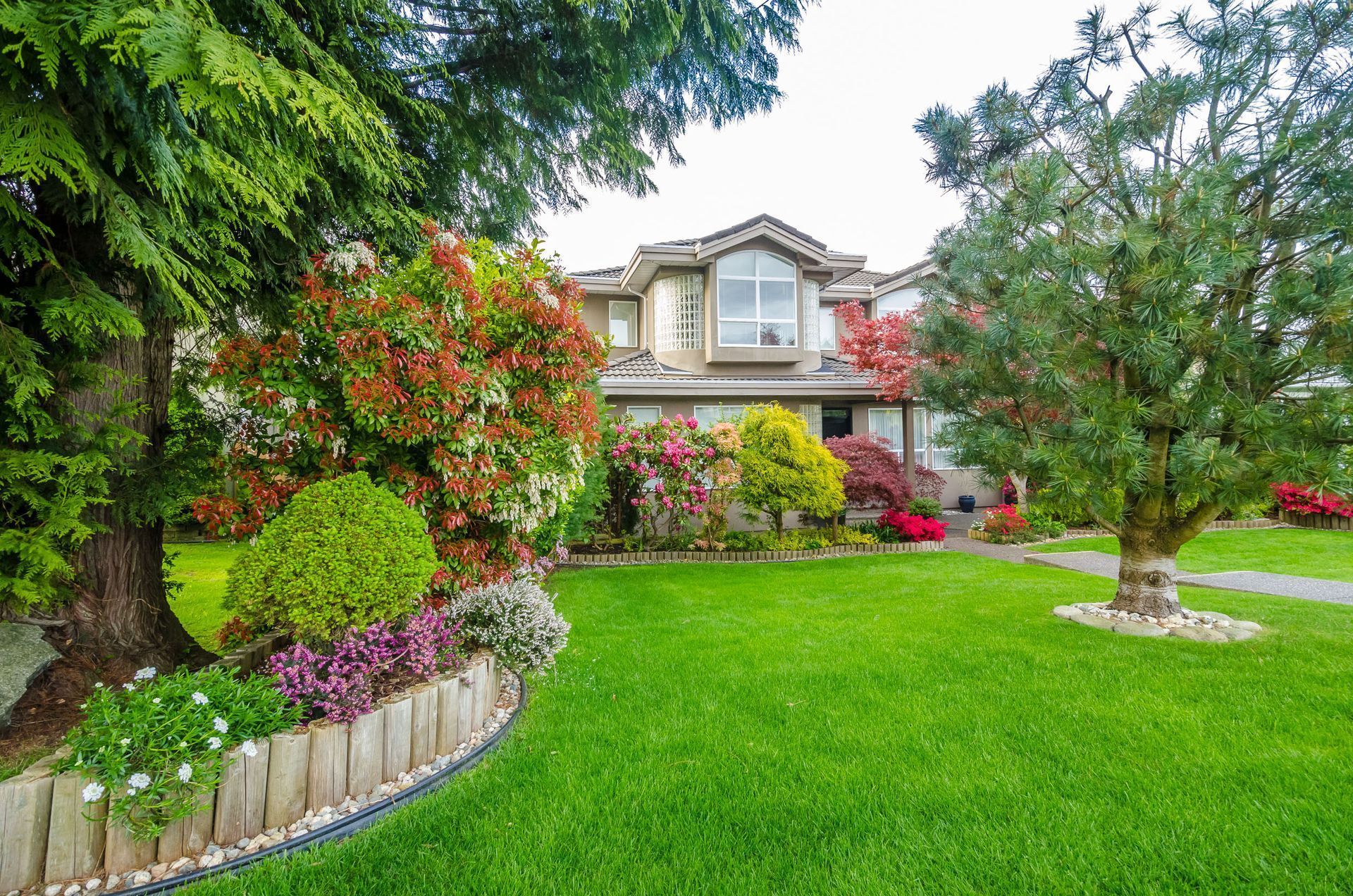 House with lush green lawn, colorful flower beds, and mature trees in the yard.