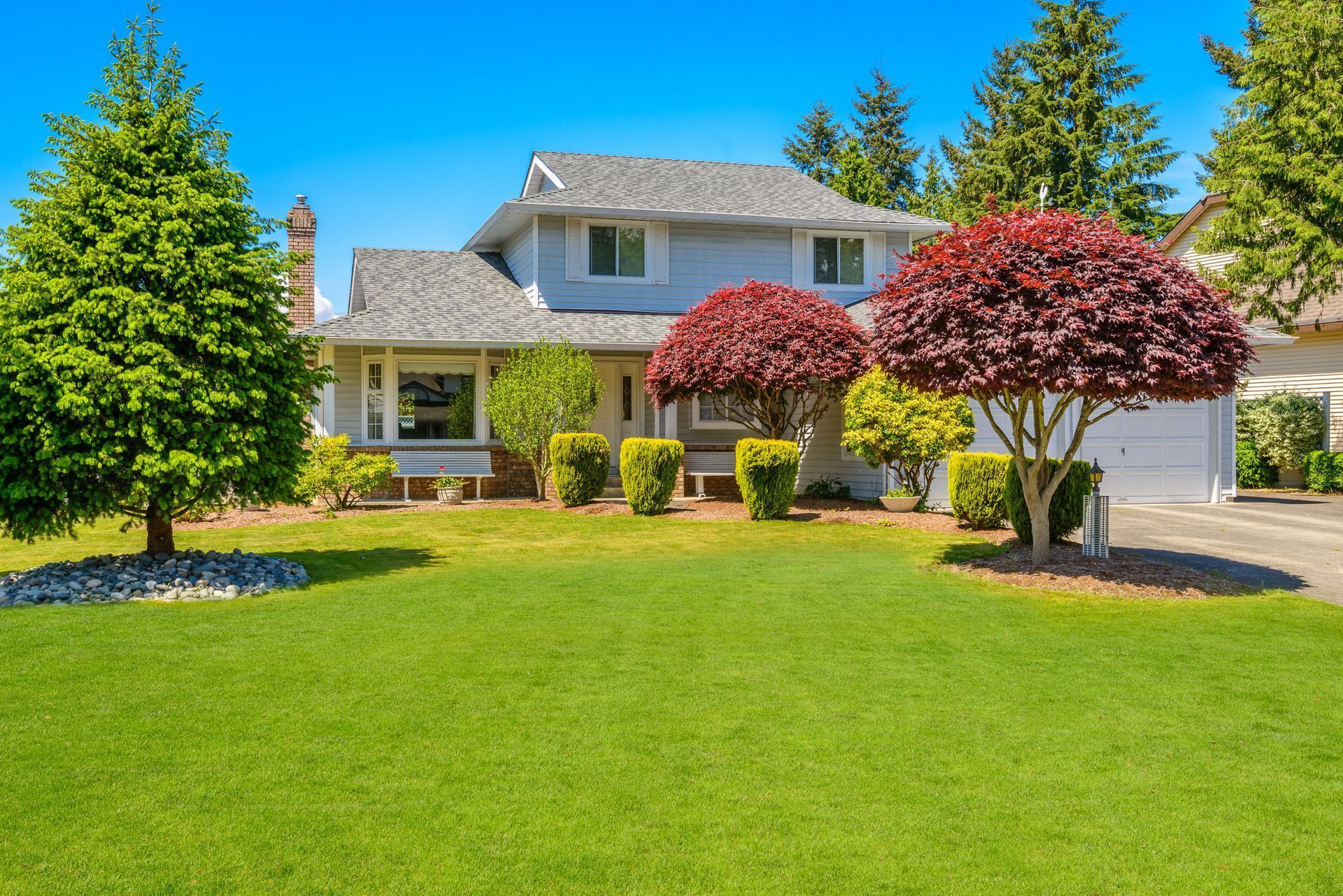 Two-story blue house with green lawn, red and green trees, and blue sky.