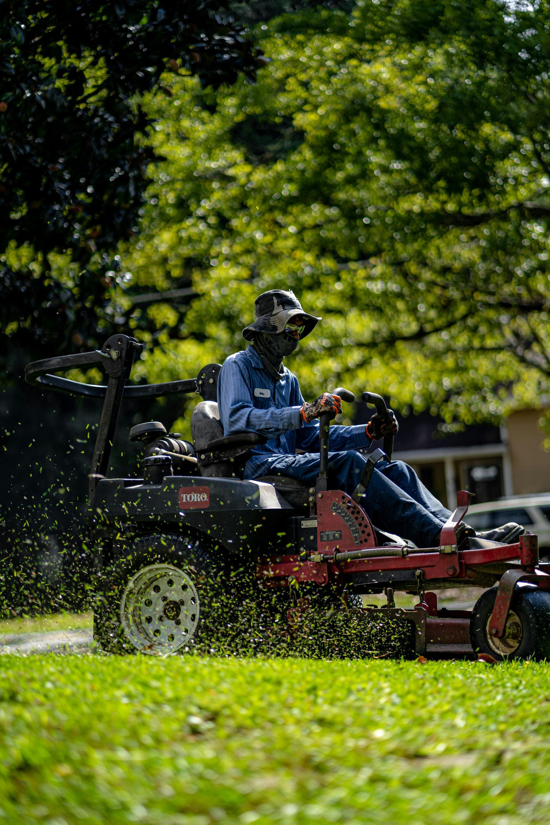 Person riding a red lawn mower through a sunlit green yard, spraying grass clippings.