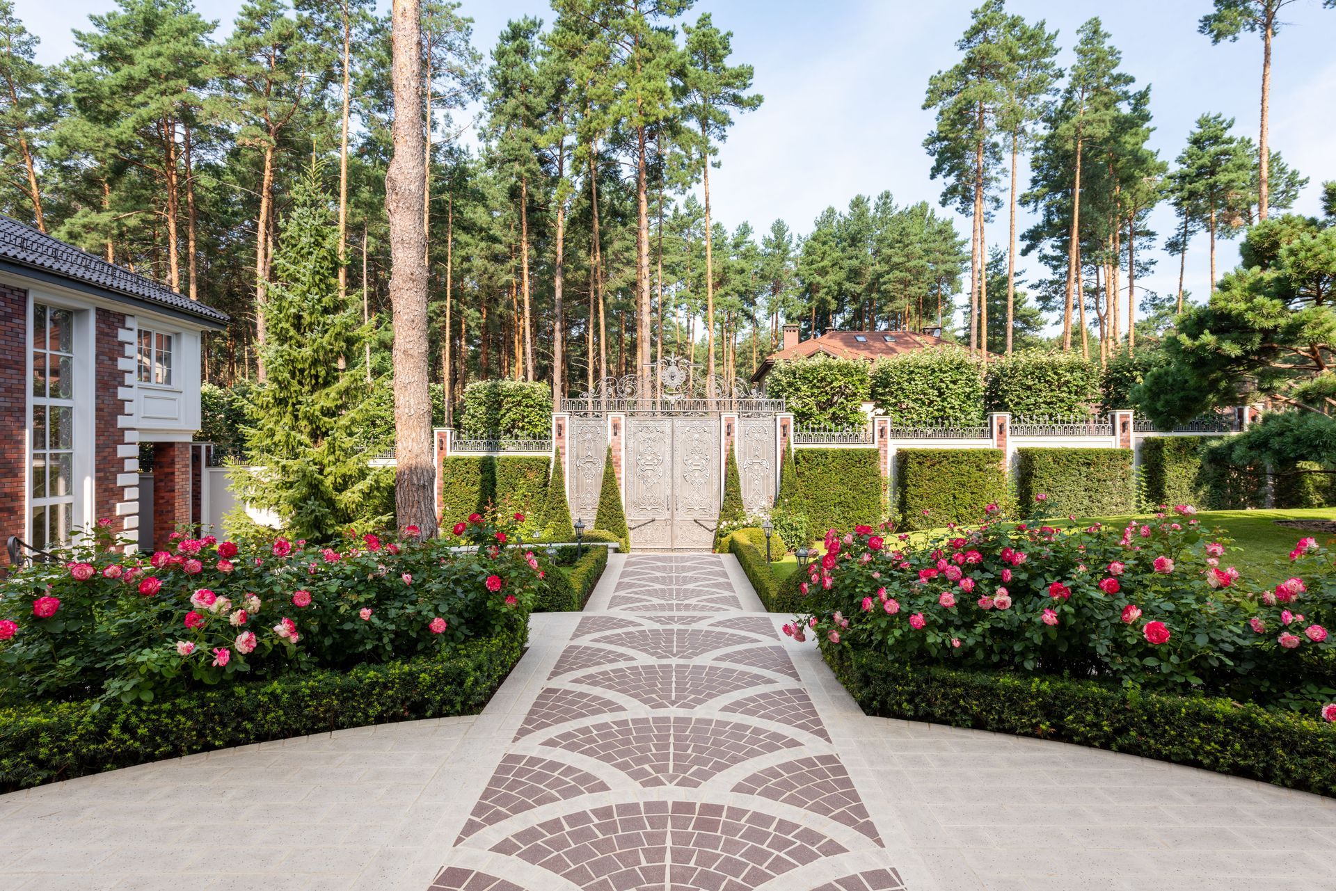 Garden path with patterned stone leading past pink flower beds, trees, and a brick house facade