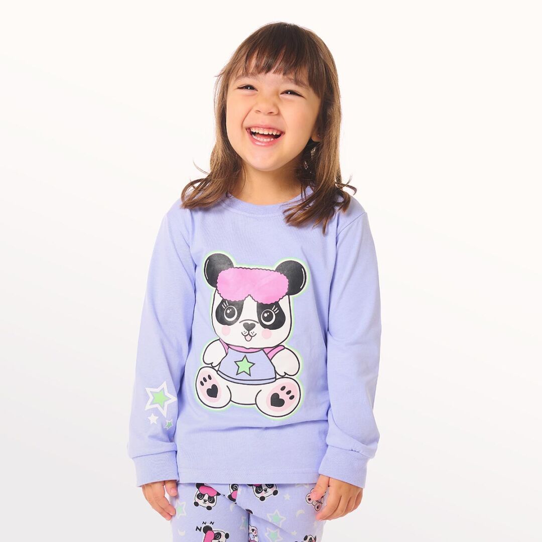 Niña pequeña posando con pijama lila junto a un gato de dibujos animados, de pie sobre un fondo blanco.