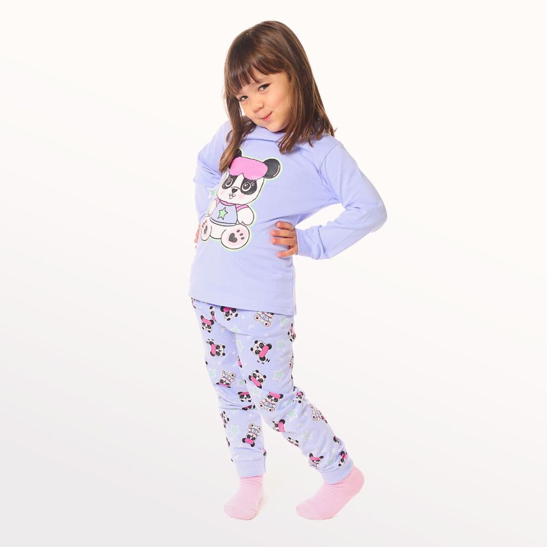 Niña pequeña posando con pijama lila junto a un gato de dibujos animados, de pie sobre un fondo blanco.