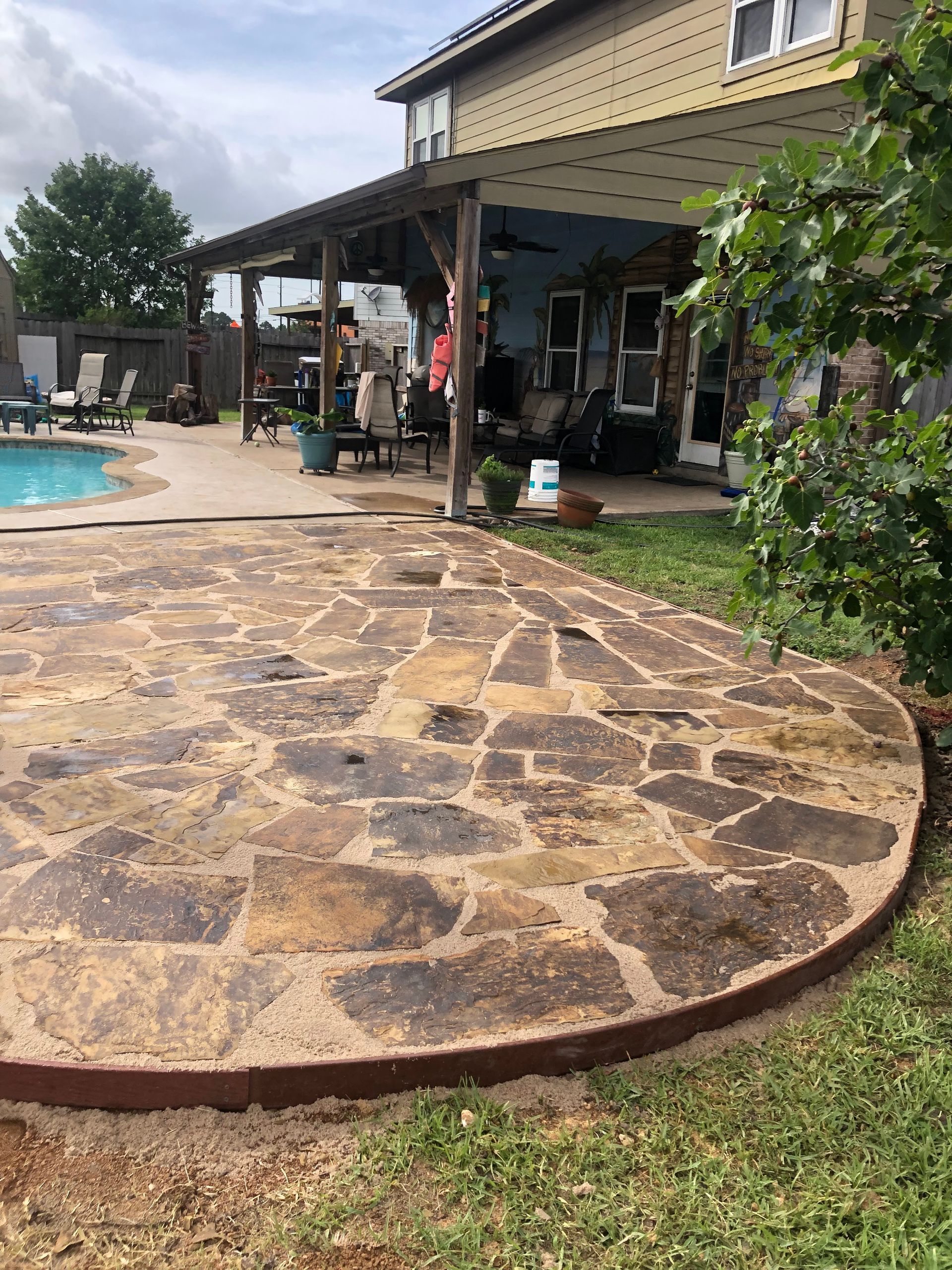 Patio with stone-like stamped concrete, pool in background, under house overhang, green grass border.