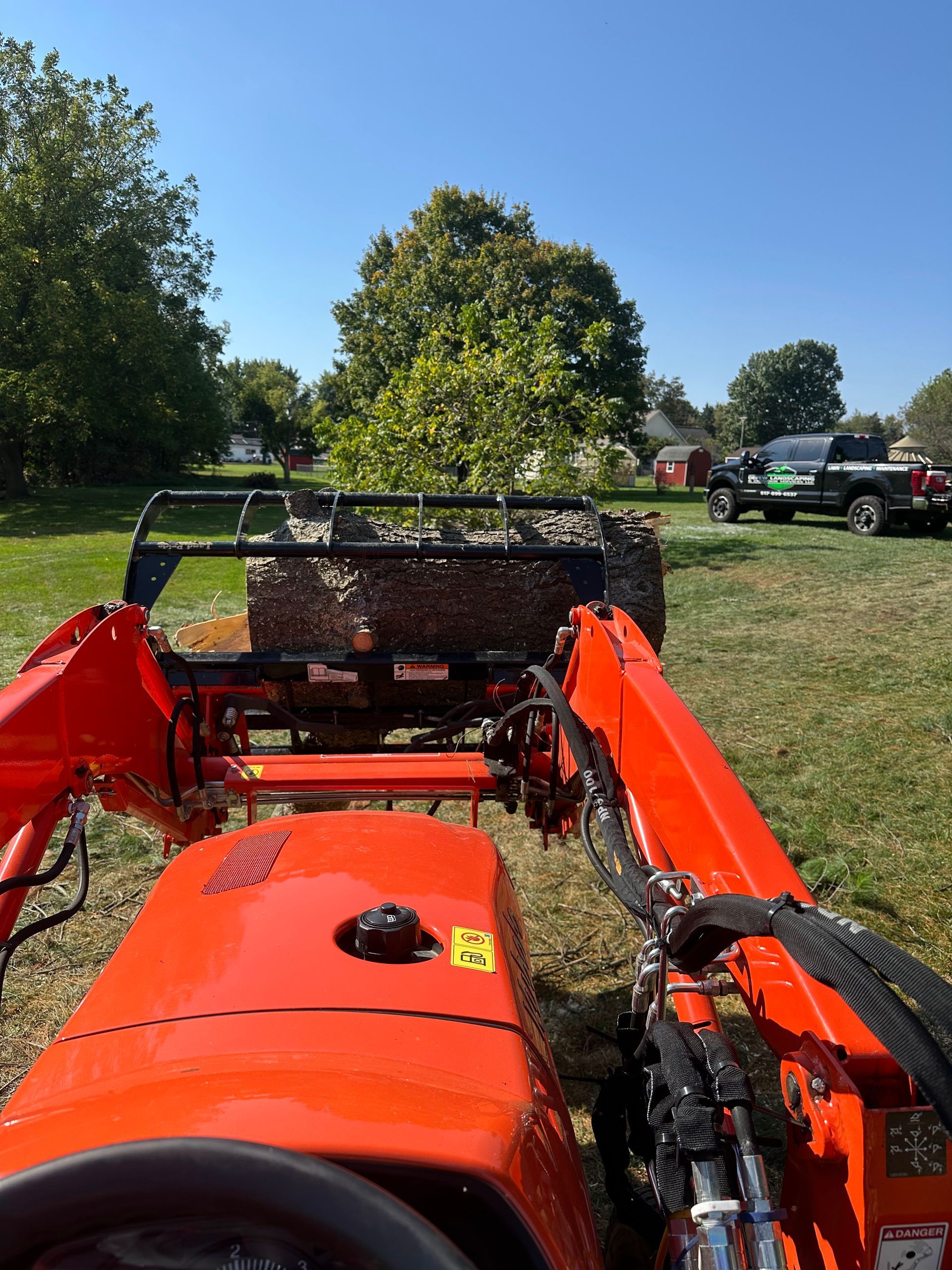 Orange tractor with loaded bucket on grass field. A truck and trees are in the background.