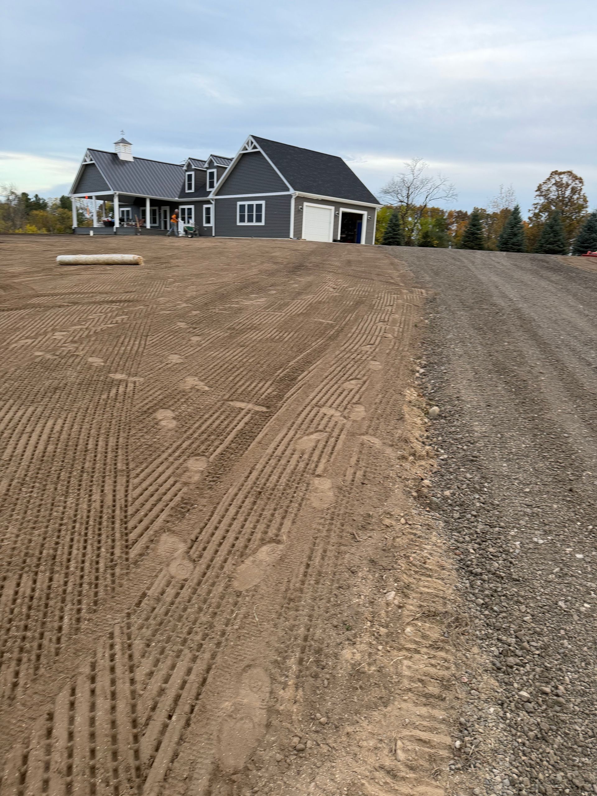 Dirt lot with a newly built house and garage, gray siding, and a gravel driveway on a cloudy day.
