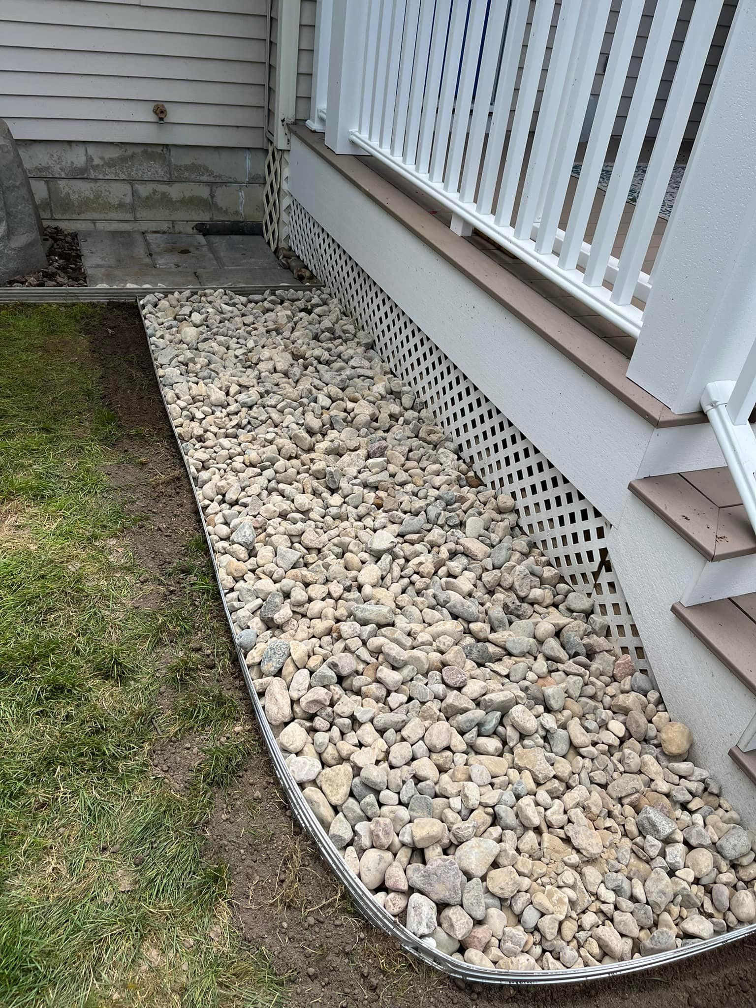 Gravel-filled bed borders a house's foundation. White lattice, stairs, and railing are visible.