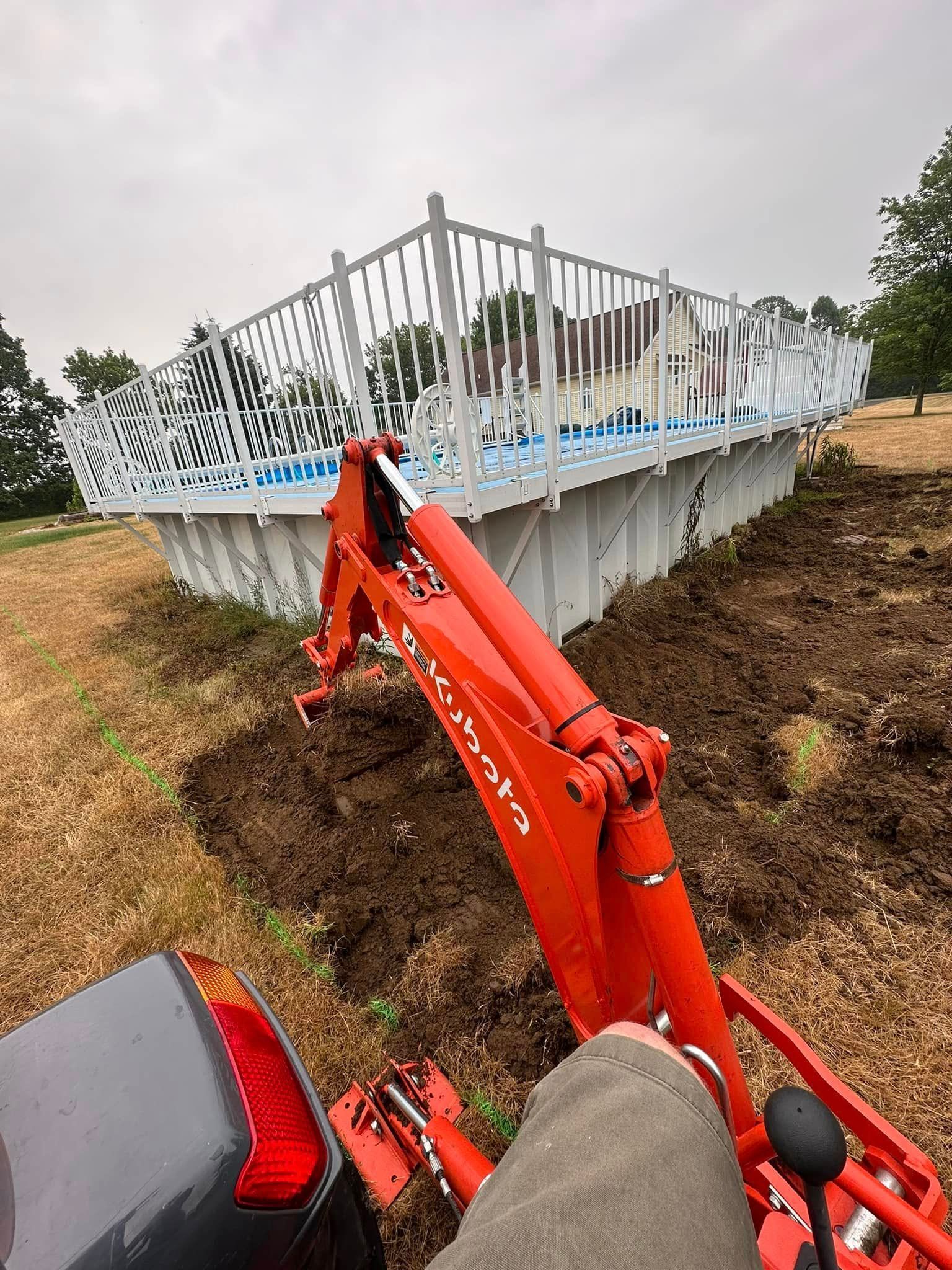 A Kubota tractor excavating dirt next to an above-ground pool with a white fence on a cloudy day.