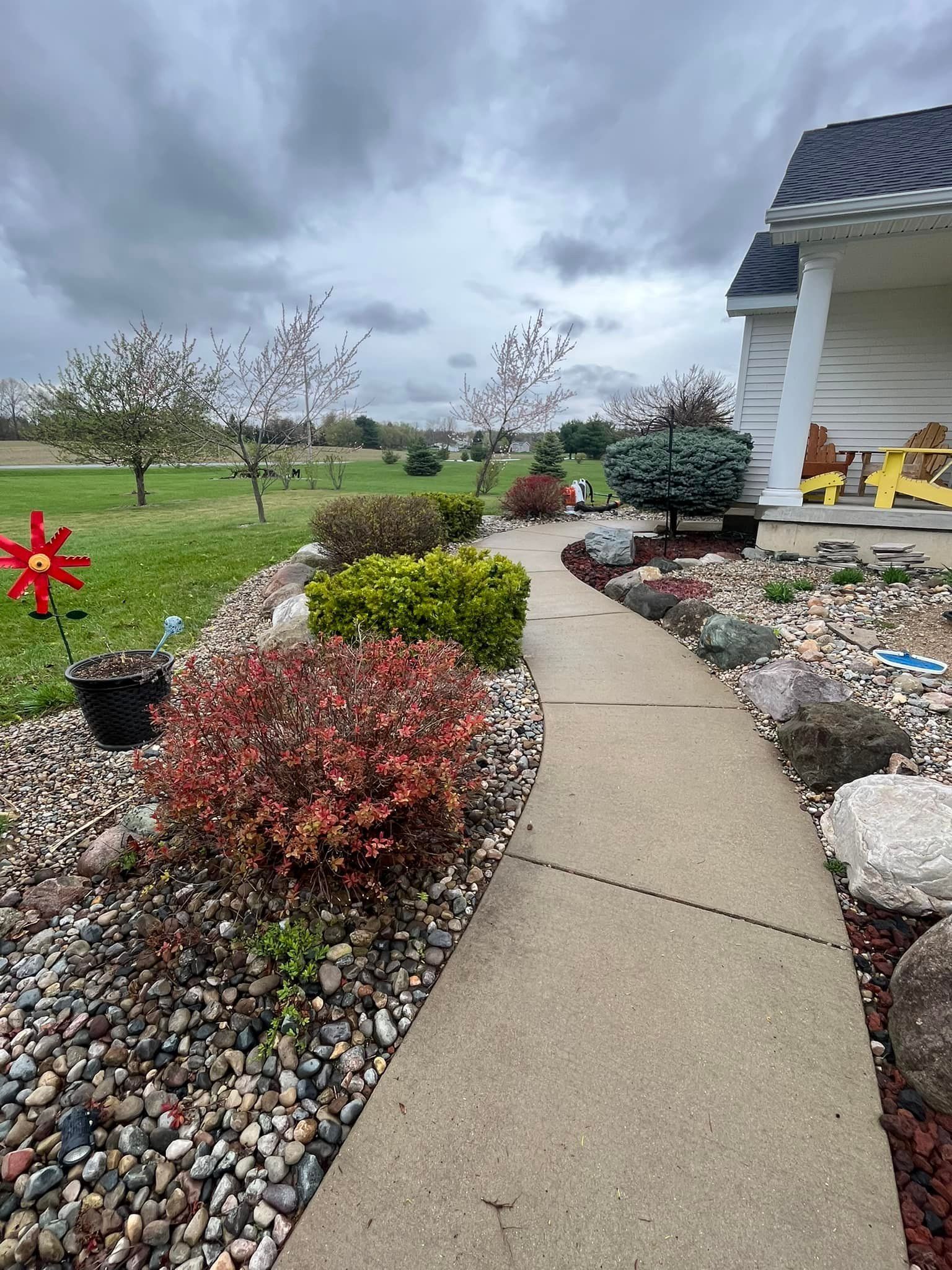 Concrete path through a landscaped garden with colorful bushes and large rocks, under a cloudy sky.