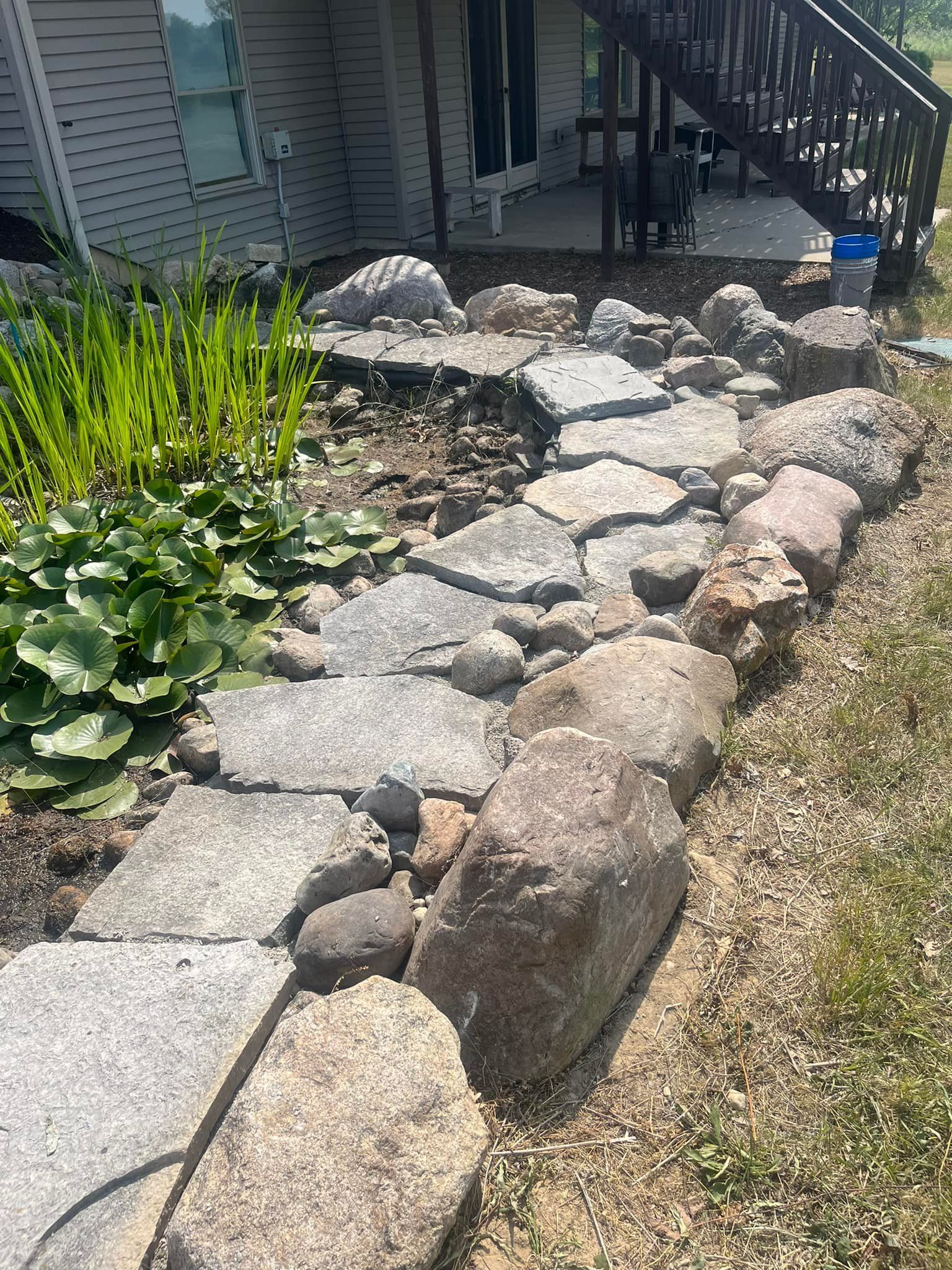 Stone path bordered by rocks and plants near a house with stairs.