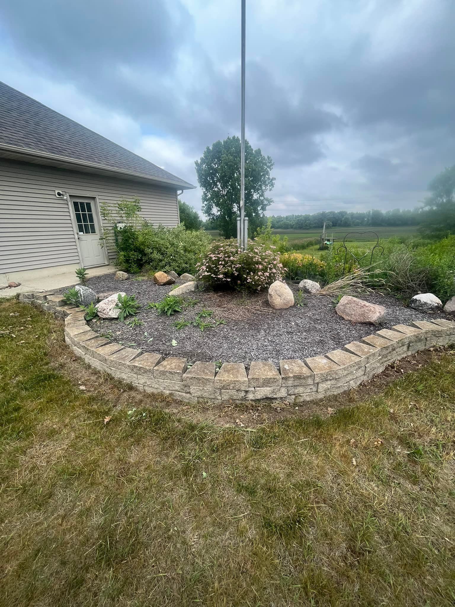 A flower bed with a stone border in front of a building and flagpole, overcast sky.
