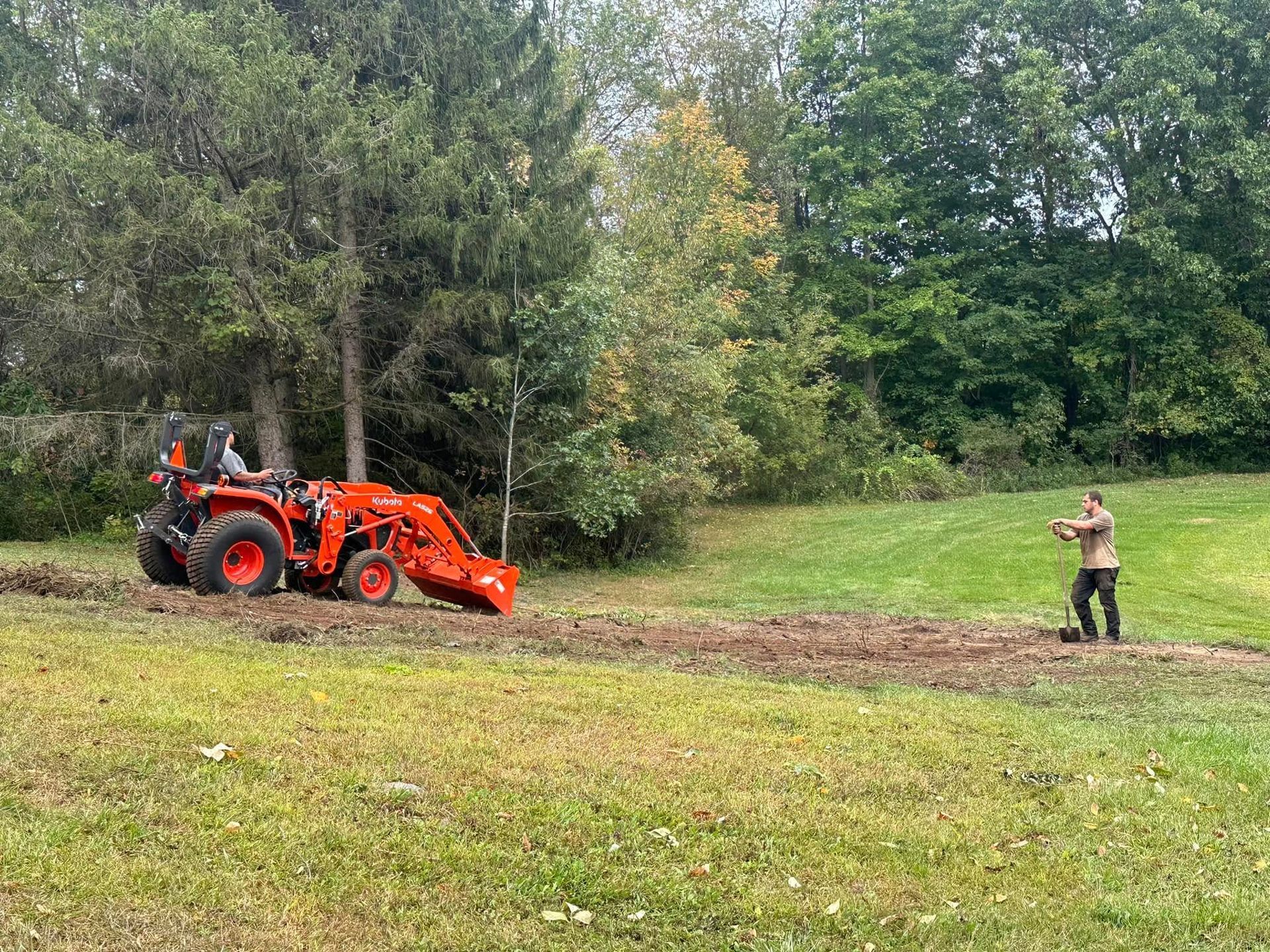 A person operating an orange tractor plowing a field, another person watches. Green trees in the background.