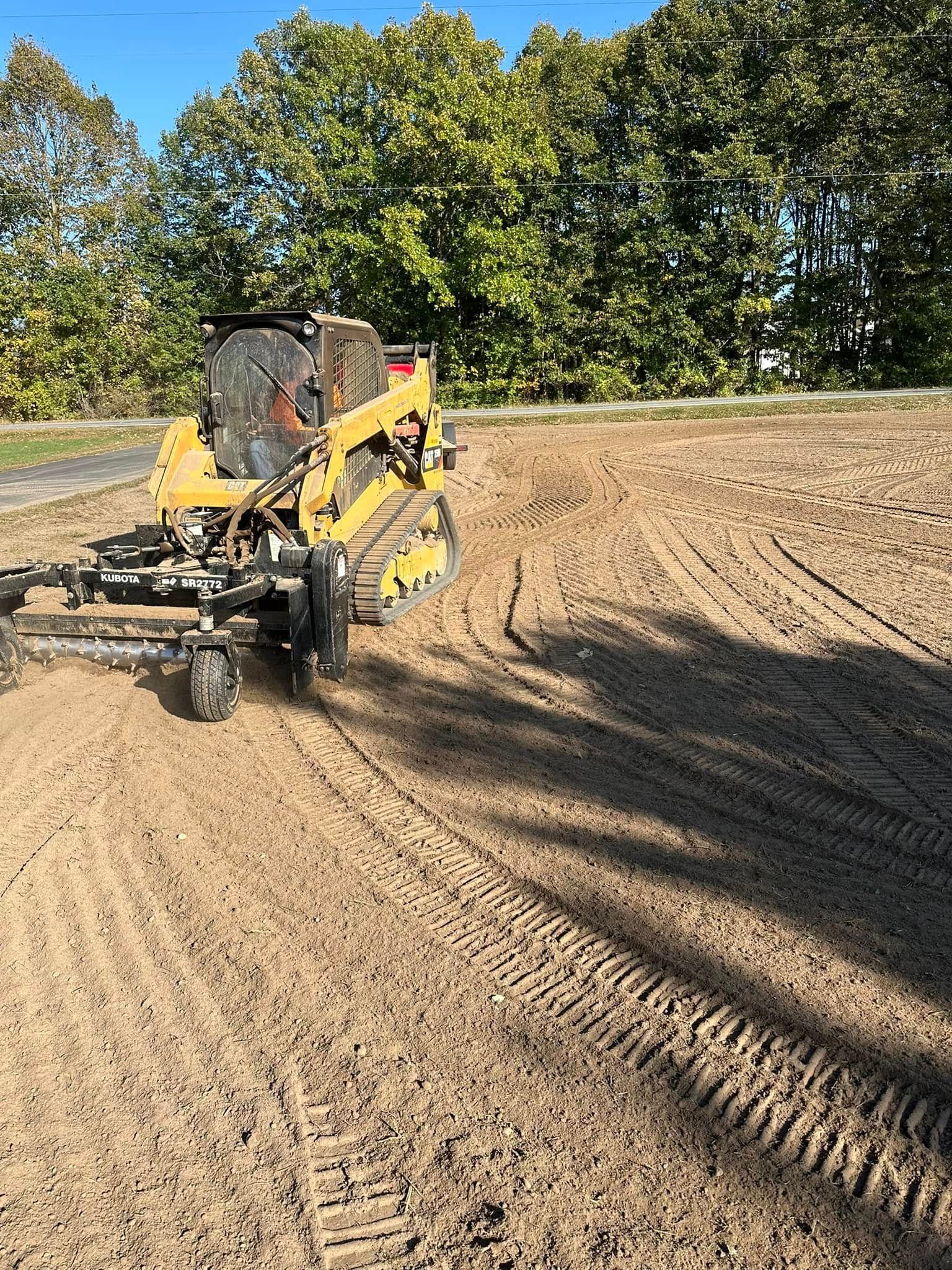 Yellow skid steer operating on a dirt field, leaving tire tracks and ground markings. Trees in background.