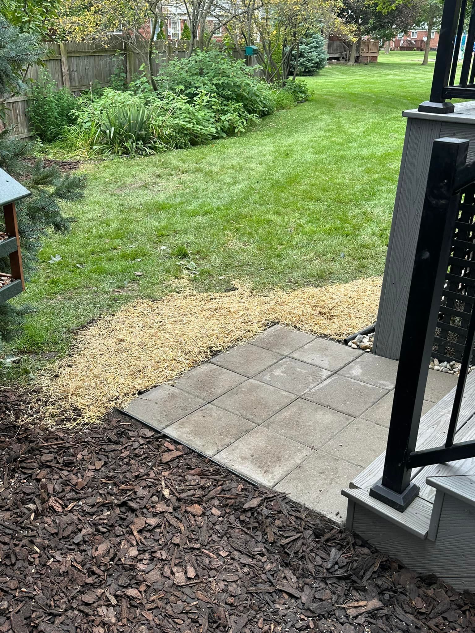 Backyard scene: concrete patio, gravel pathway, grassy lawn, mulch, and a deck with black railing.
