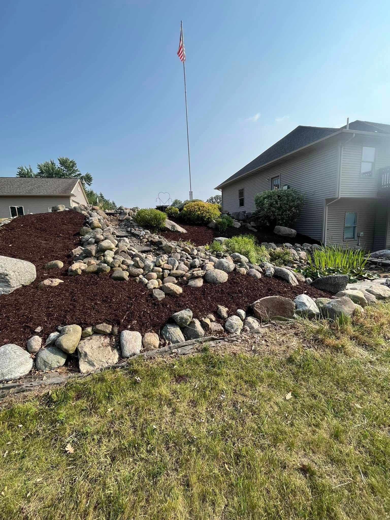 A hillside landscape with rocks, mulch, and a house under a clear sky. A flag pole stands tall.