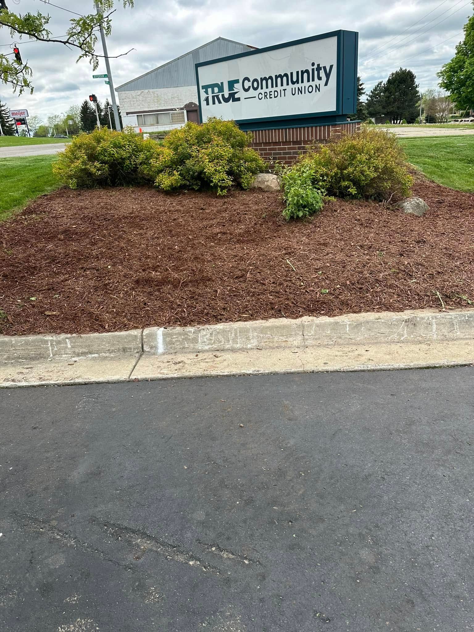 Sign for True Community, brown mulch bed with green bushes, asphalt road.