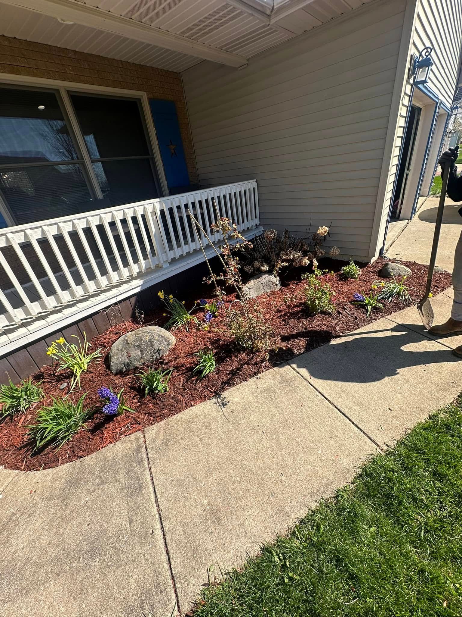 Front yard garden bed with red mulch, plants, and large rocks next to a white porch railing and a concrete walkway.