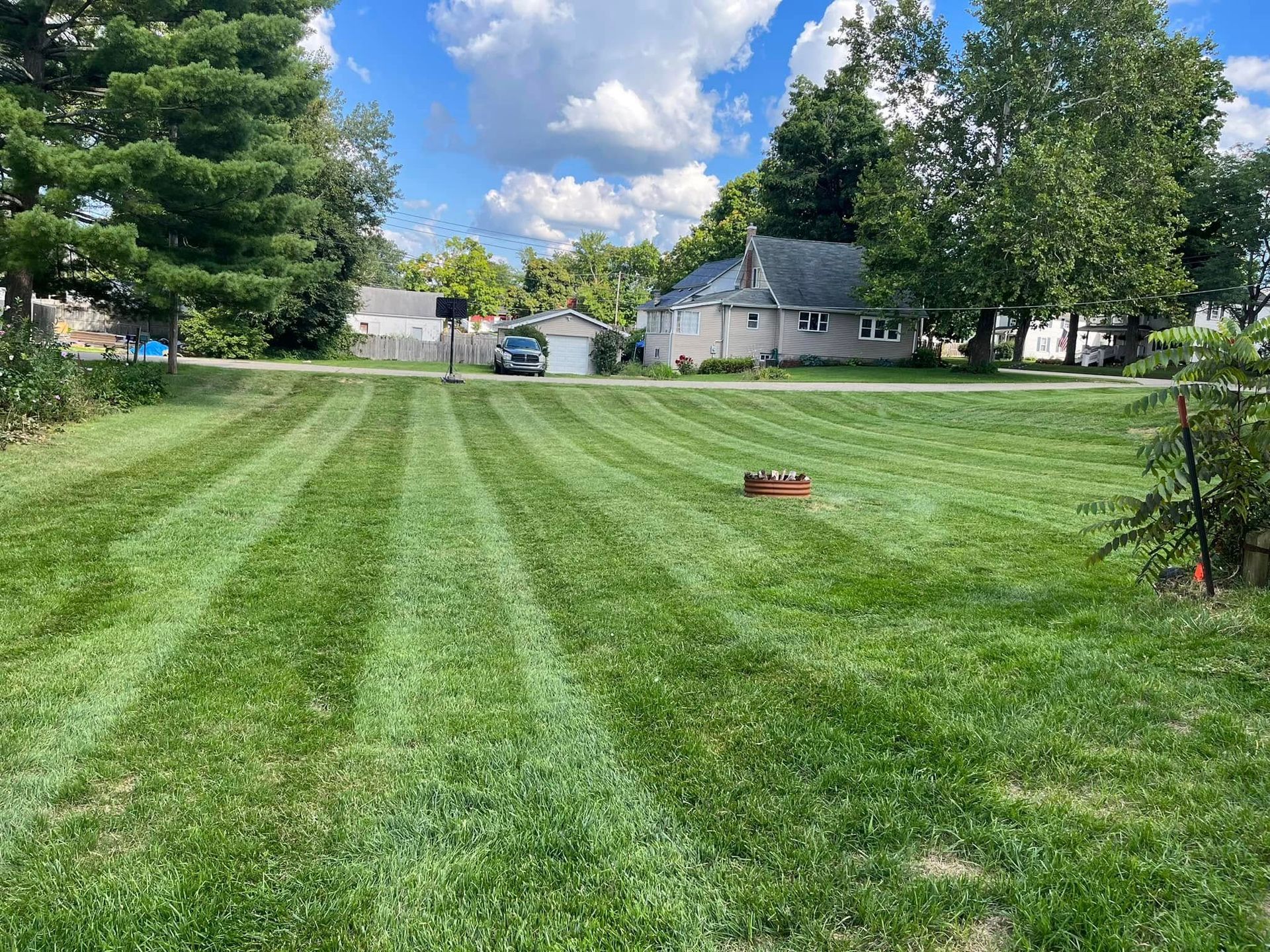 Lawn with mowed stripes, small house in the background, a car, and a blue sky.