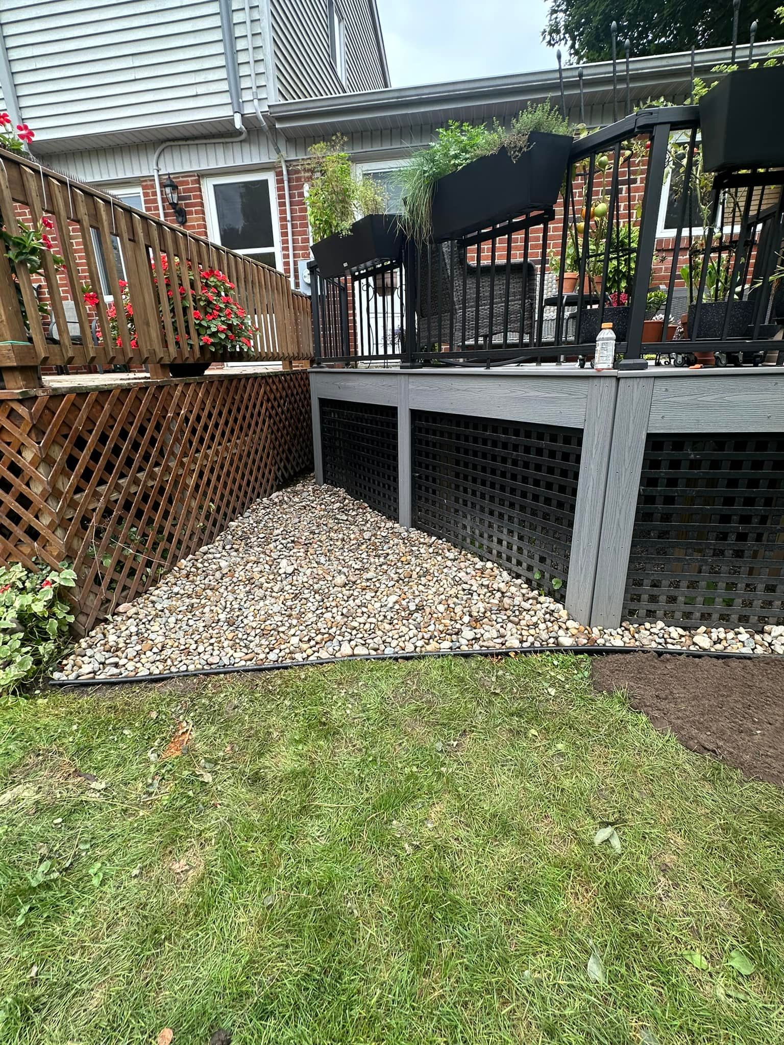 Deck with lattice, gravel base, and green grass. House in the background with plants and a cloudy sky.